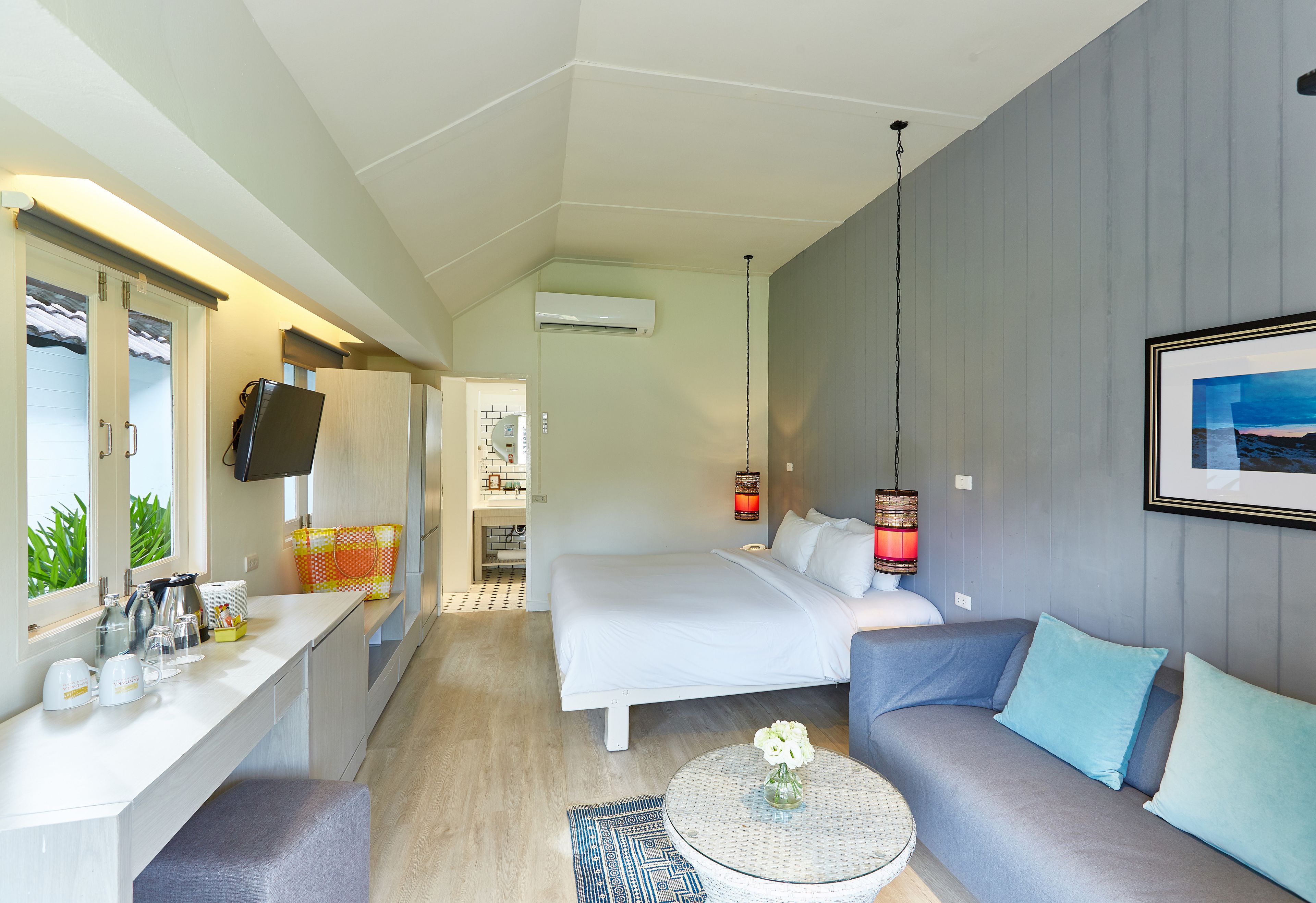 A compact hotel room with a bed, a sofa, a small table with a vase, a kitchen area, and a TV.