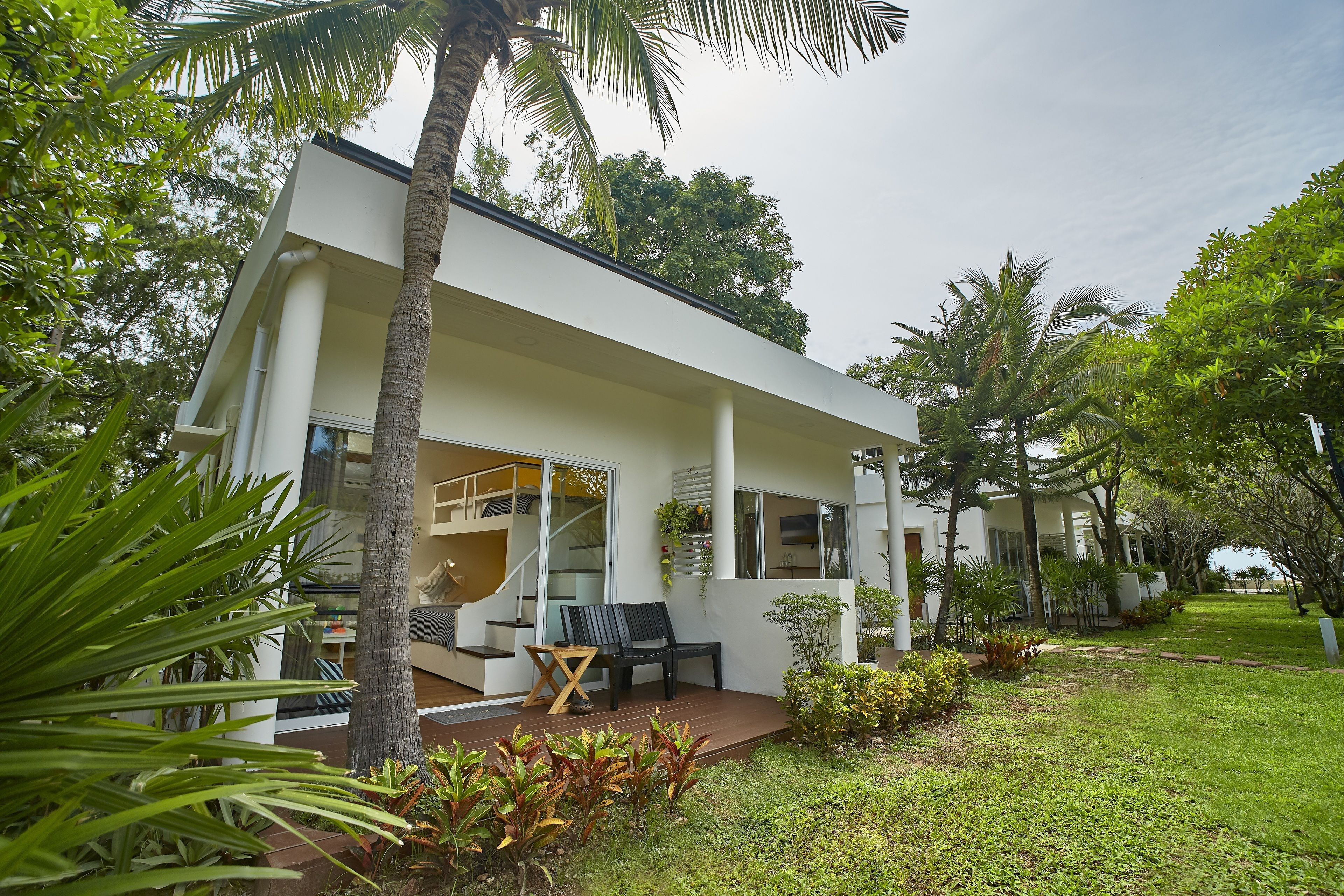 A modern house with a covered porch, a staircase leading to an upper level, and a garden with palm trees and shrubs.