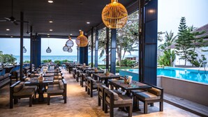 Un restaurante frente al mar con terraza, ventanas amplias y lámparas de mimbre colgantes.