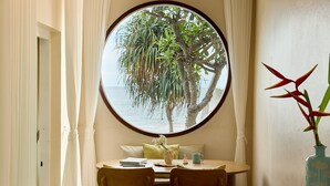 Una habitación con ventanal redondo que da a un árbol y al agua, una mesa de comedor con sillas y un florero con una planta.