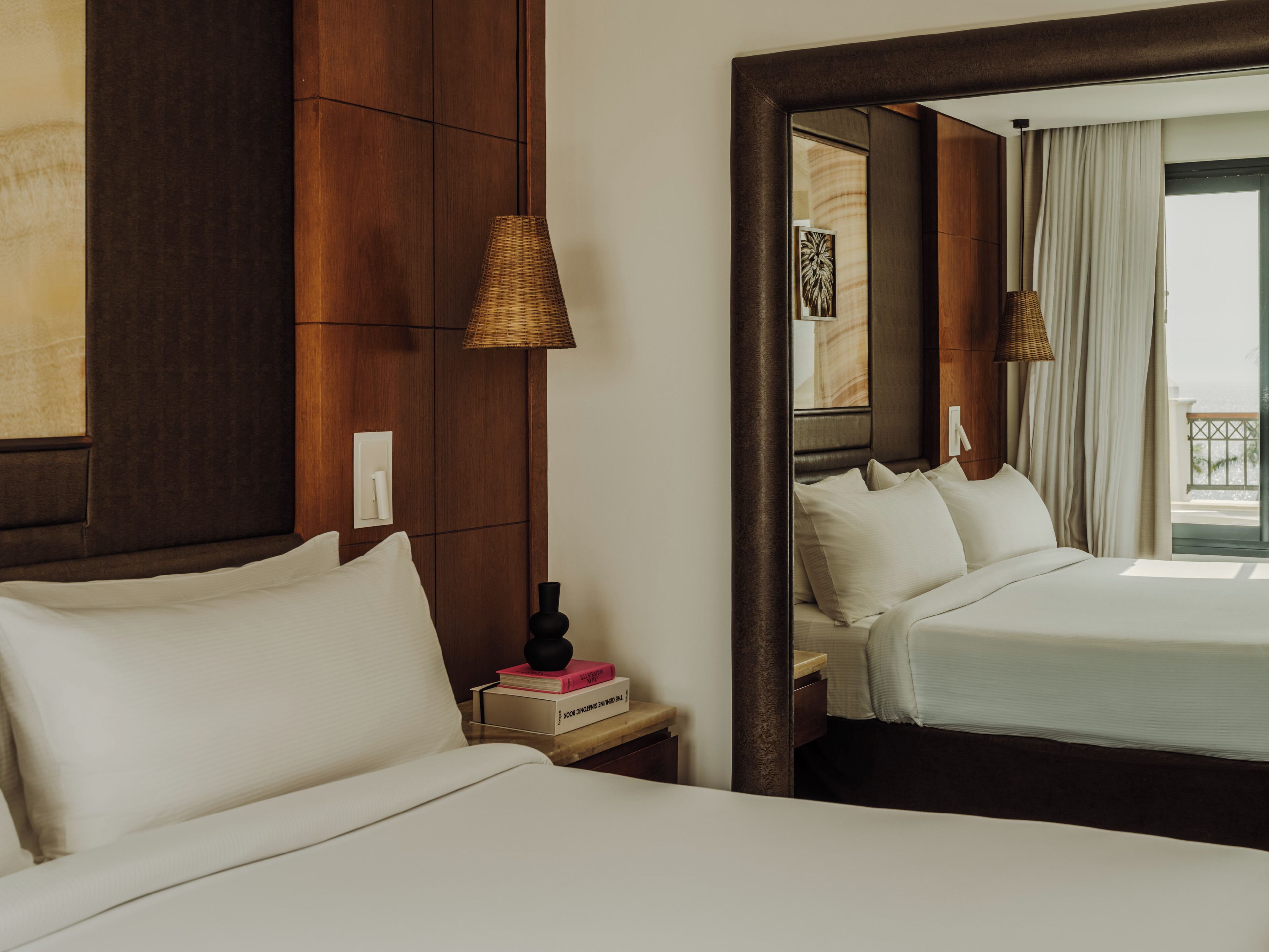 Habitación de hotel con dos camas, una mesita de noche con lámpara, un espejo y vistas al exterior.