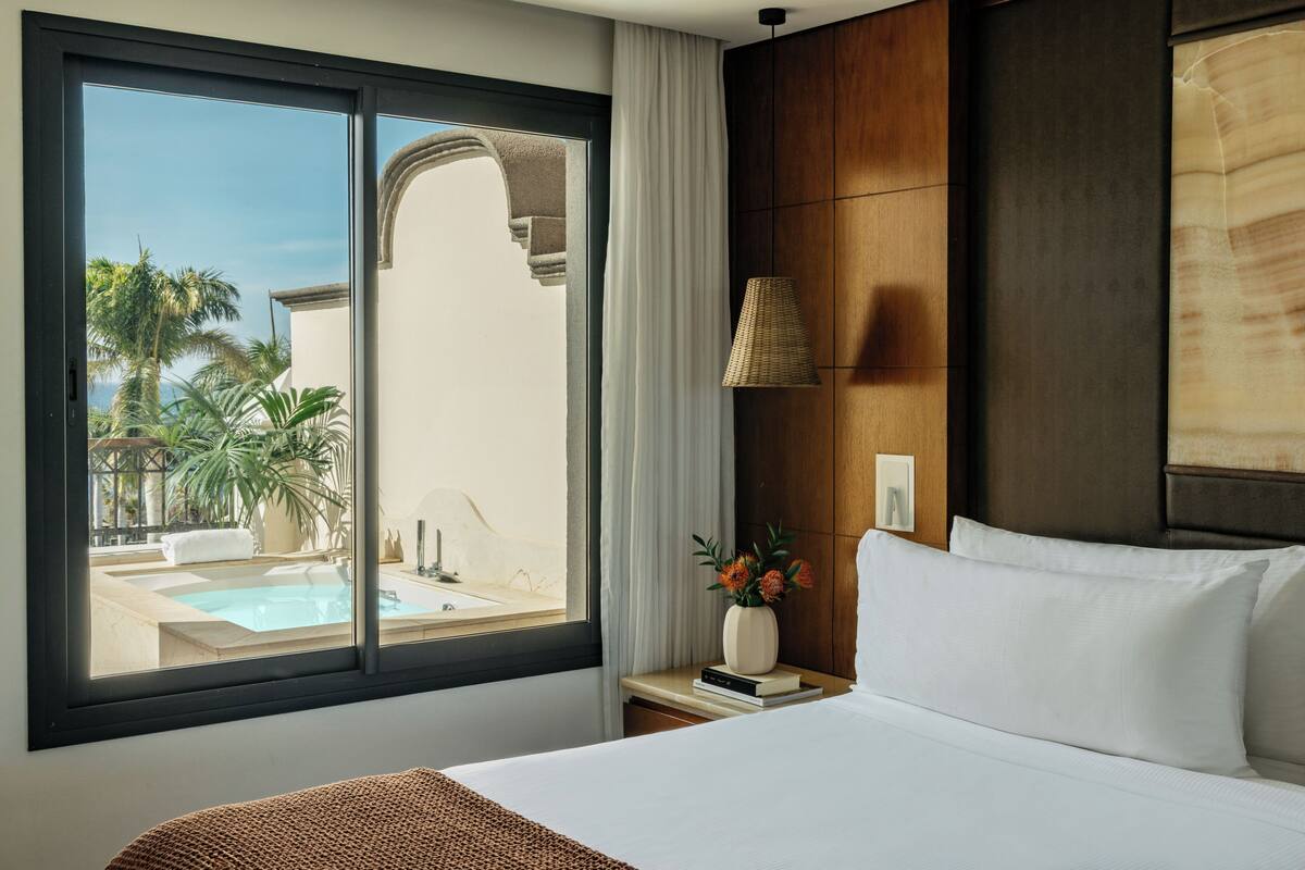 Ein Hotelzimmer mit großem Fenster, einem Bett, einem Nachttisch mit Blumenvase und Blick auf Pool und Palmen.