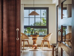 An open doorway leading to a room with a dining table, two chairs, a hanging light fixture, and a window with a view of palm trees.