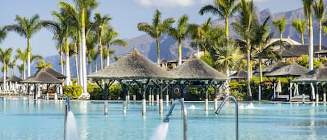 Una piscina con fuentes de agua, rodeada de palmeras y estructuras de techo de paja.