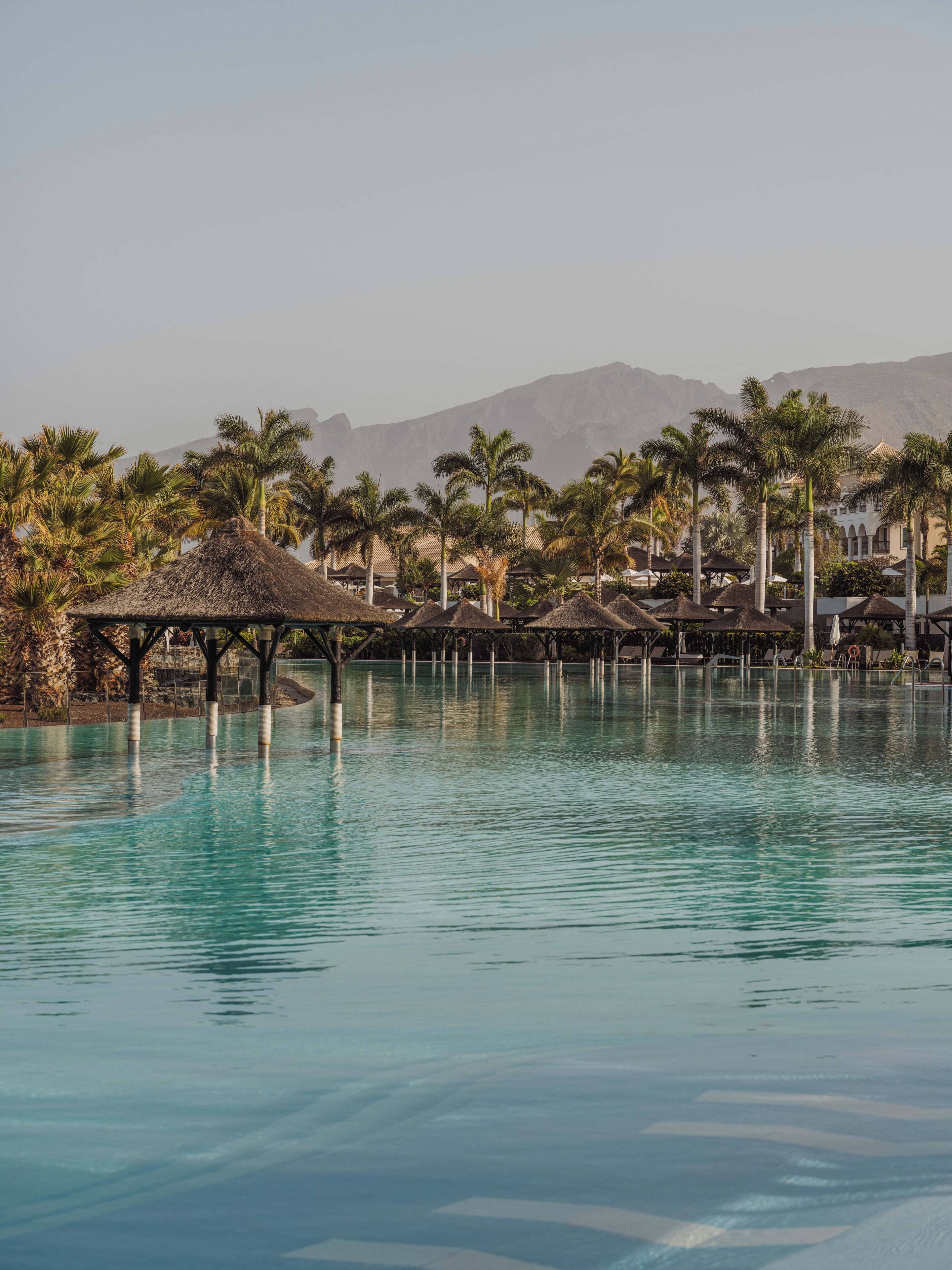 Zona de piscina en un resort, con palmeras y estructuras de techo de paja.