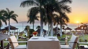 An outdoor dining area with a table set for two, overlooking a beach with palm trees and a sunset. - RedLevel at Gran Melia Palacio de Isora - Adults Only (Guia de Isora)