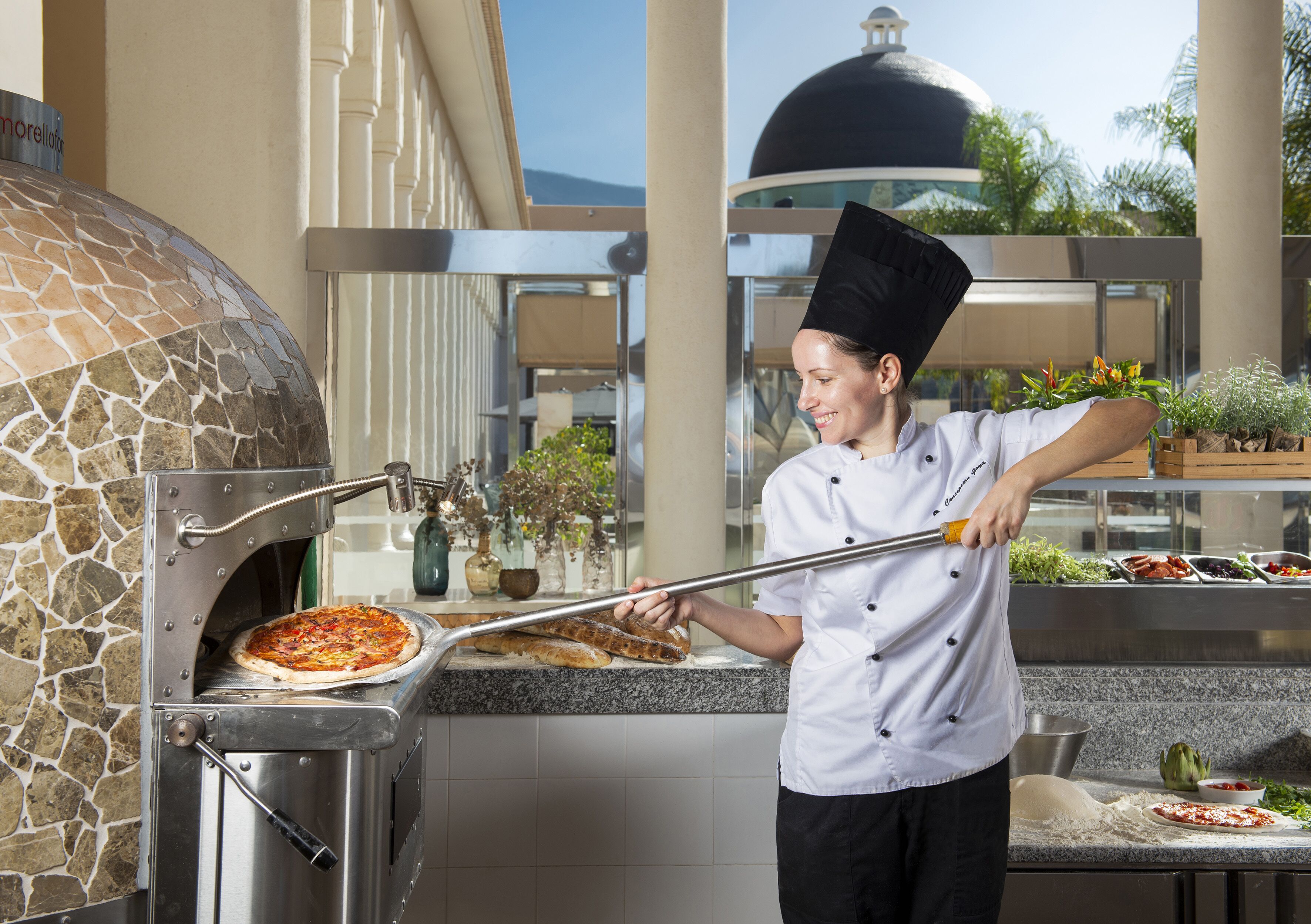 Un cocinero prepara pizza en un horno de piedra, con vistas a un edificio y plantas al fondo.