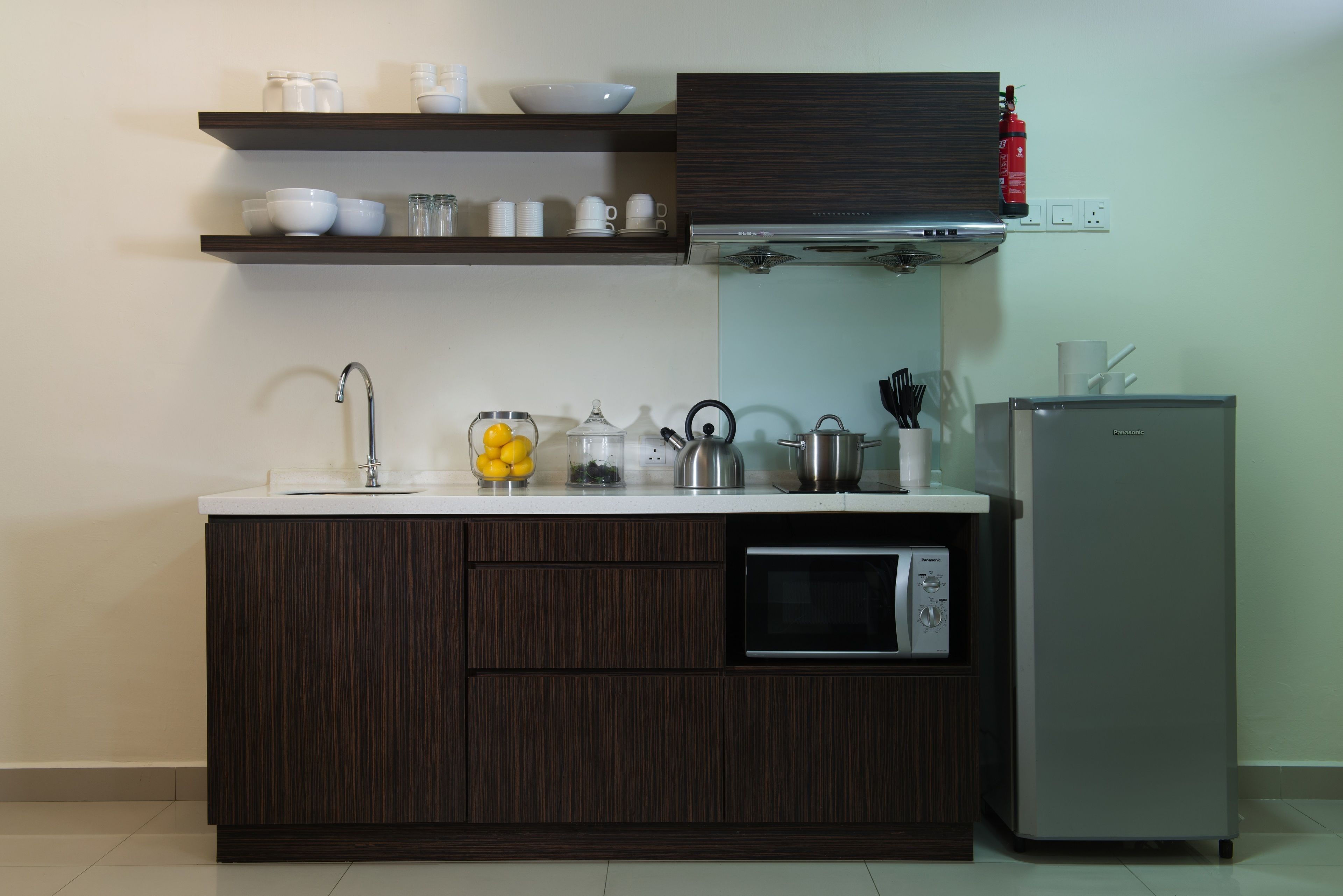 A modern kitchen with dark wood cabinets, a white countertop, and a built-in microwave. There are shelves above the counter holding dishes and glasses.