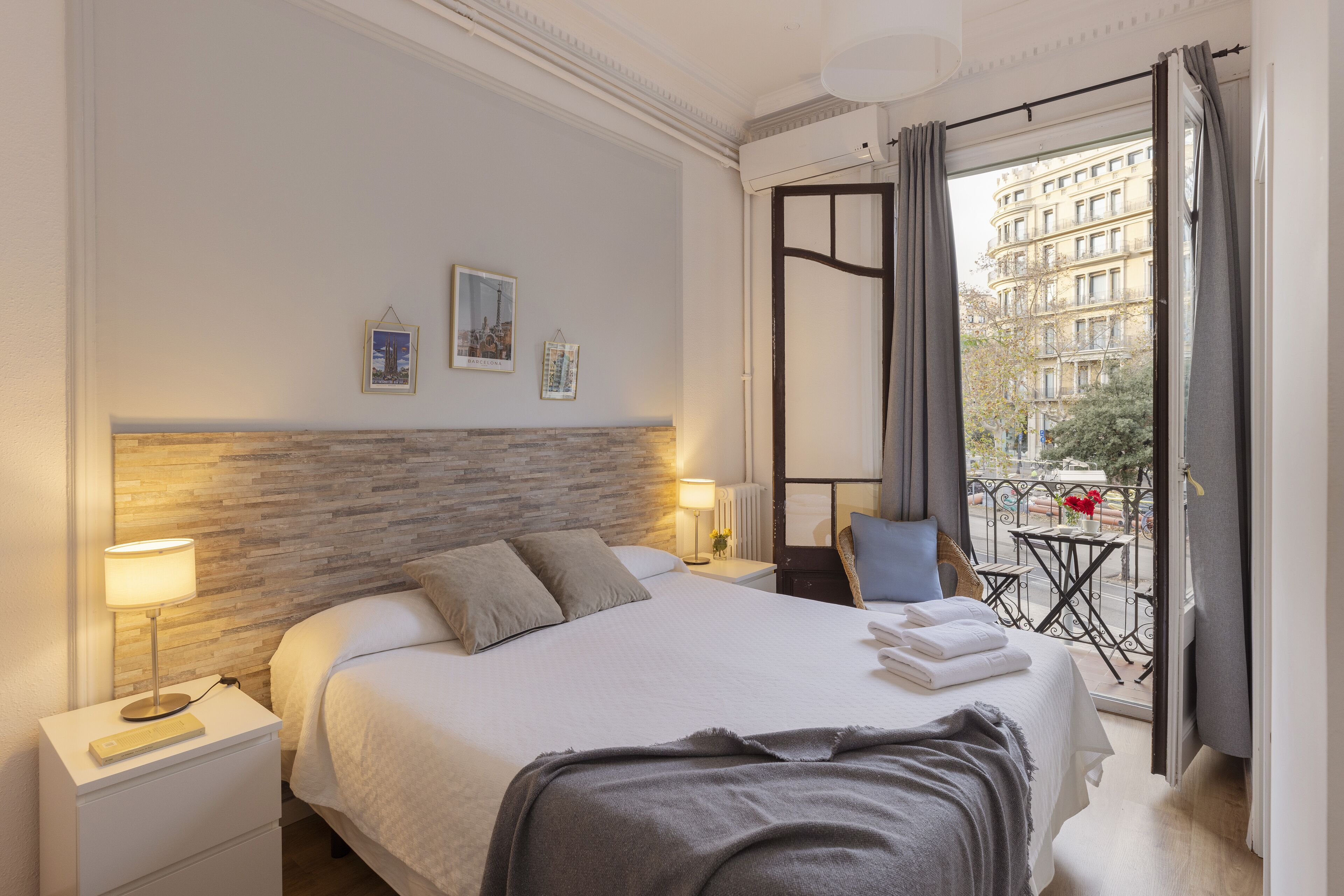 Habitación de hotel con una cama bien hecha, una mesita de noche con lámpara, vistas a la ciudad a través de la ventana y un balcón con mesa y sillas.