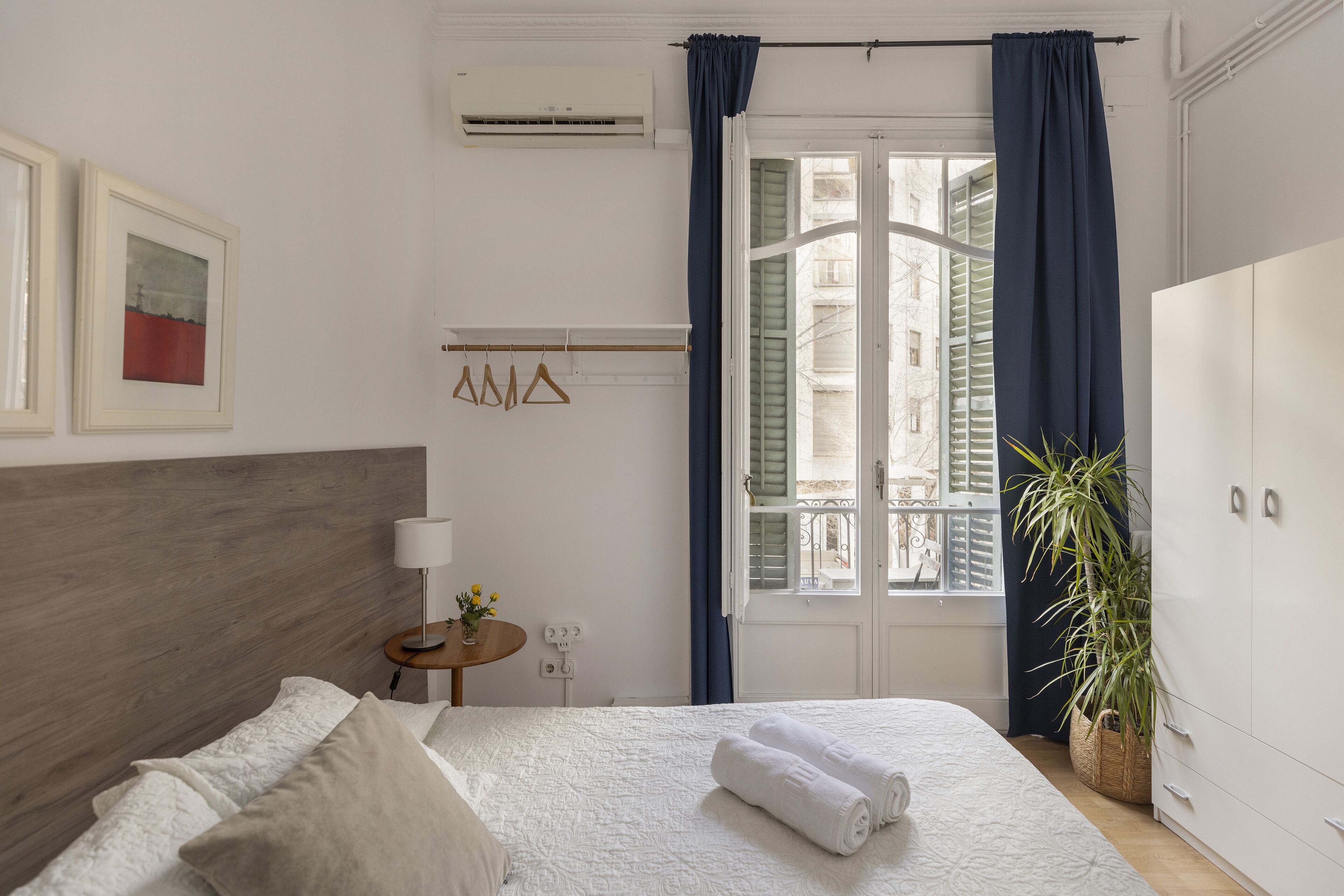 A neatly arranged hotel room with a bed, a bedside table with a lamp, a small plant, and a wardrobe.
