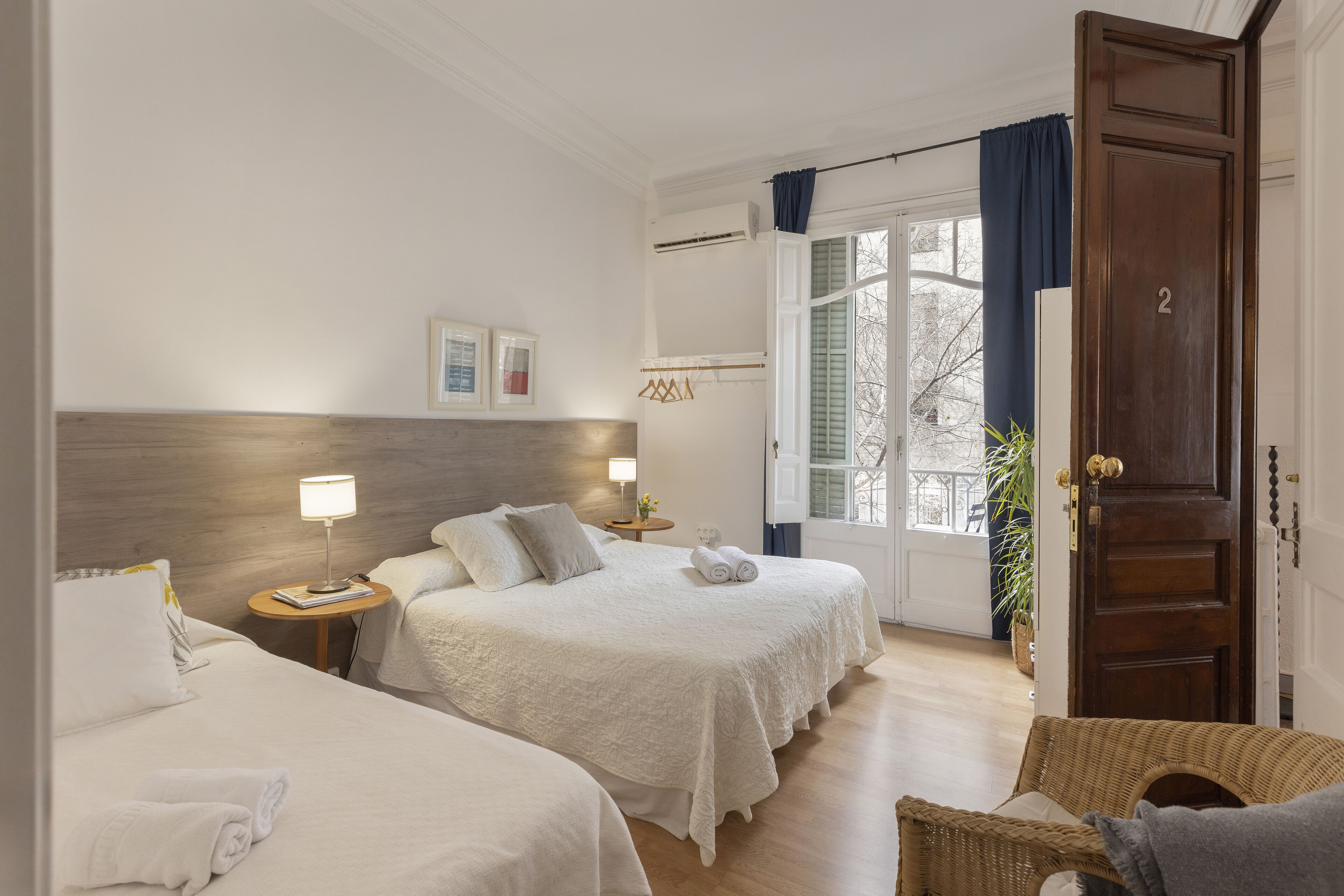 Quarto de hotel com duas camas, cabeceira de madeira, luminárias de cabeceira, uma cadeira de vime e vista para o exterior.