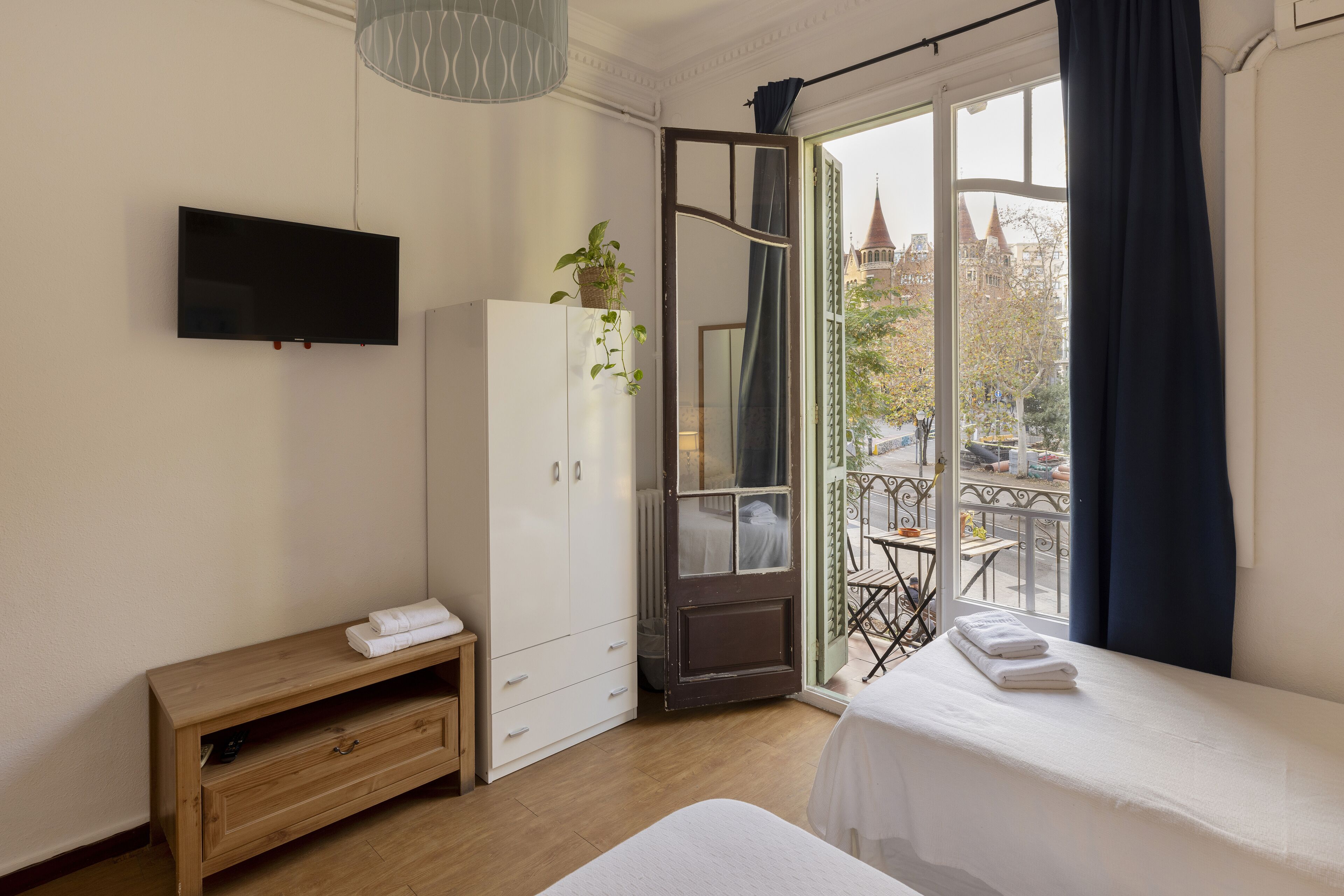 Habitación de hotel con cama, televisor montado en la pared, armario, una pequeña mesa de madera y balcón con mesa y sillas.