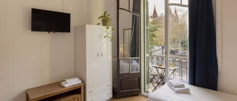 Habitación de hotel con cama, televisor montado en la pared, armario, una pequeña mesa de madera y balcón con mesa y sillas.