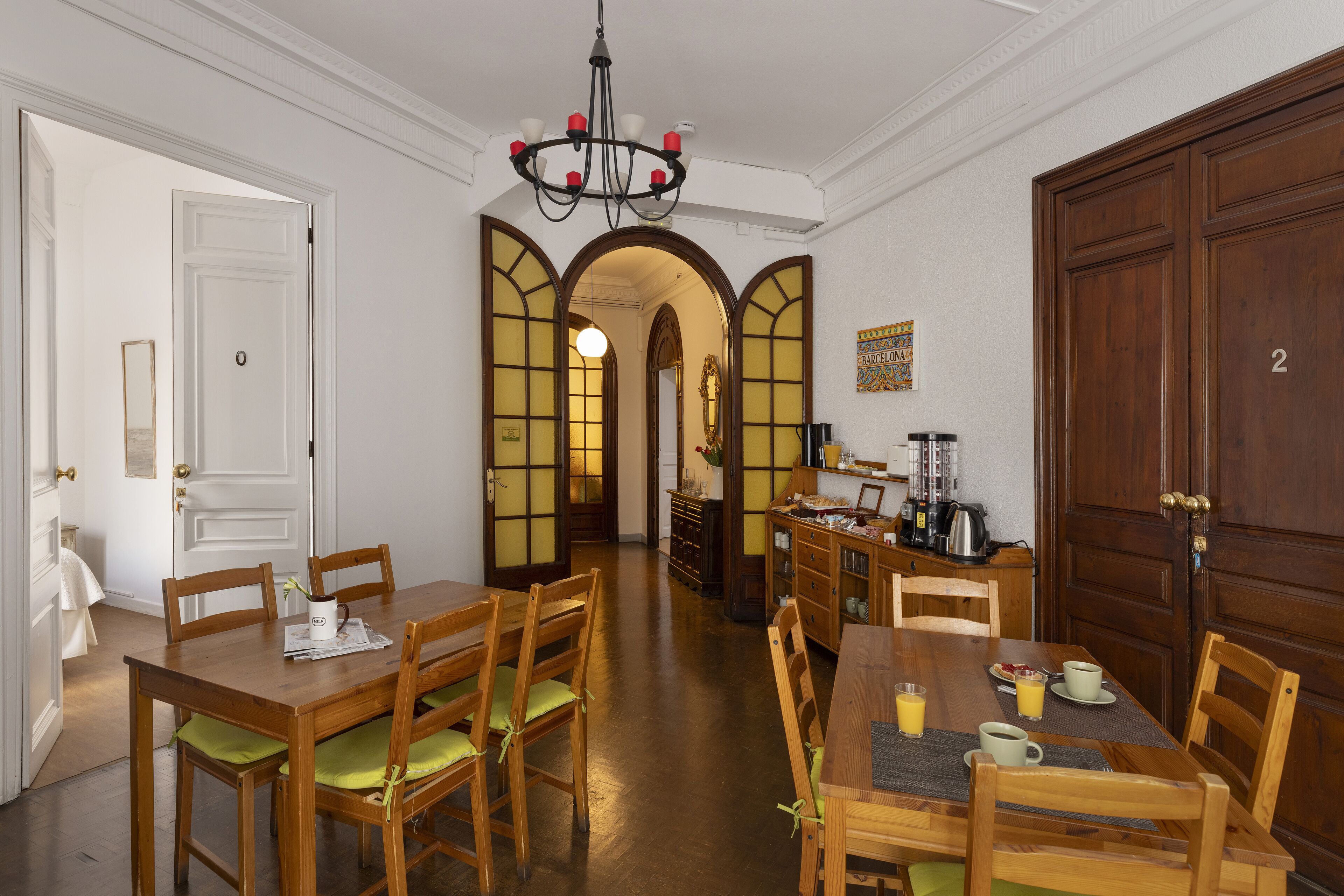 A dining area with wooden tables and chairs, a chandelier, and a kitchen area with a coffee machine and cabinets.