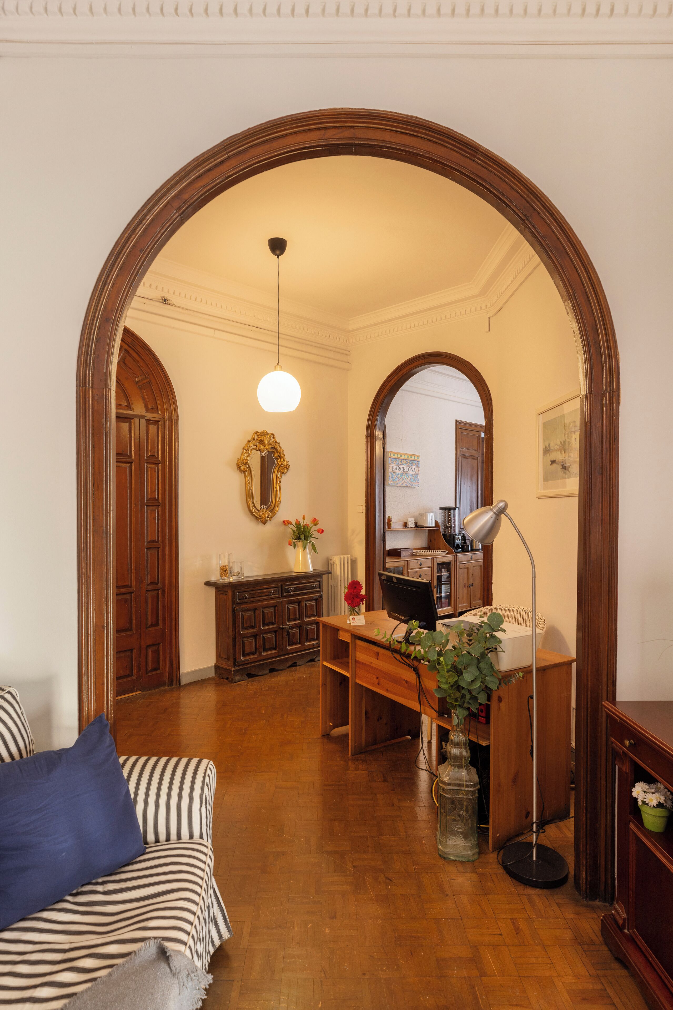 Una habitación con suelo de madera, un sofá a rayas, un escritorio con lámpara, un espejo y un amplio arco de entrada.