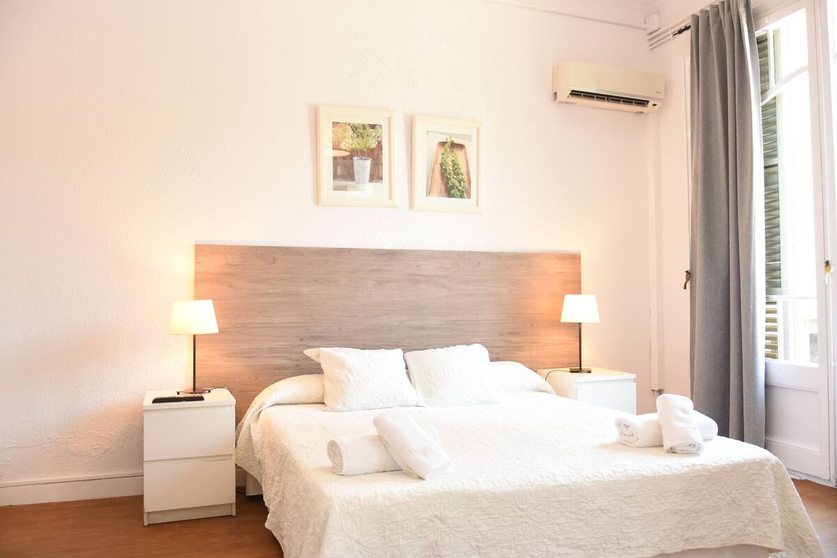 Hotellihuone, jossa on puinen sängynpääty, kaksi yöpöytää valaisimineen, valkoisilla vuodevaatteilla varustettu sänky sekä kaksi kehystettyä kuvaa seinällä.