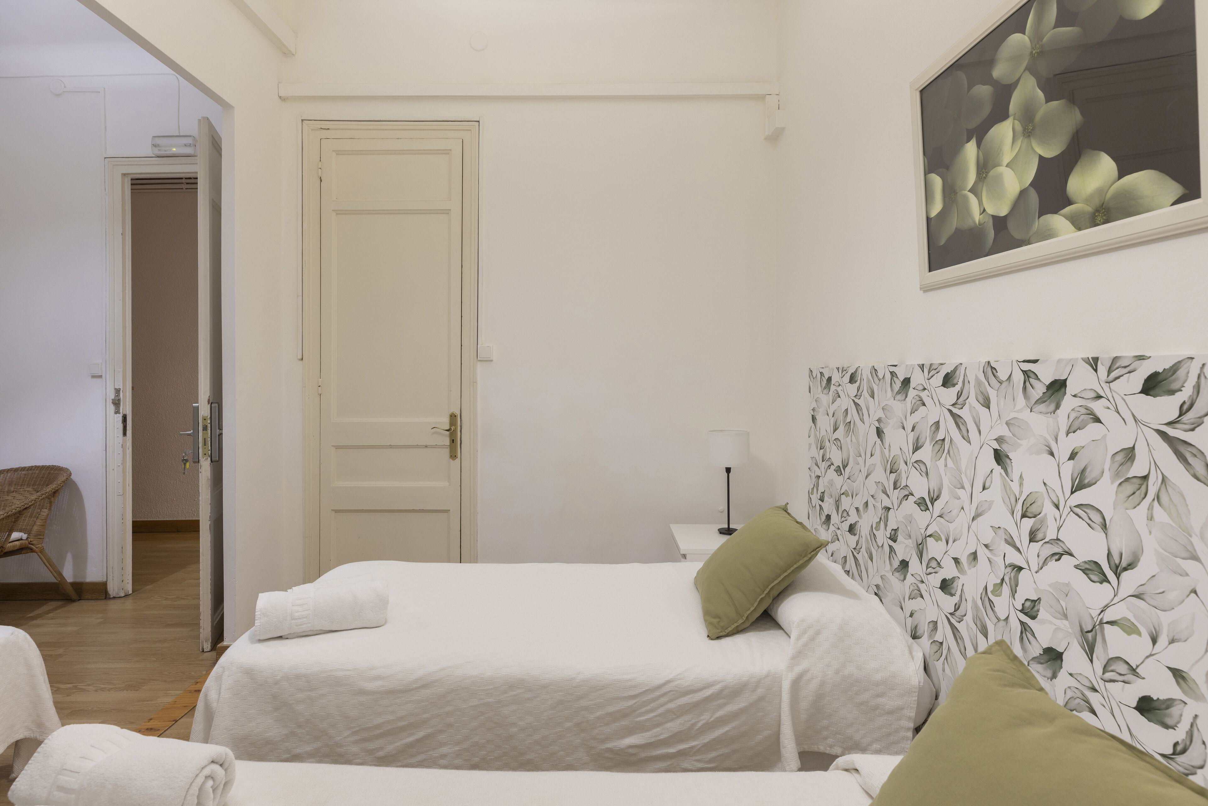 A hotel room with two beds, a door, a wall-mounted floral artwork, and a wicker chair.