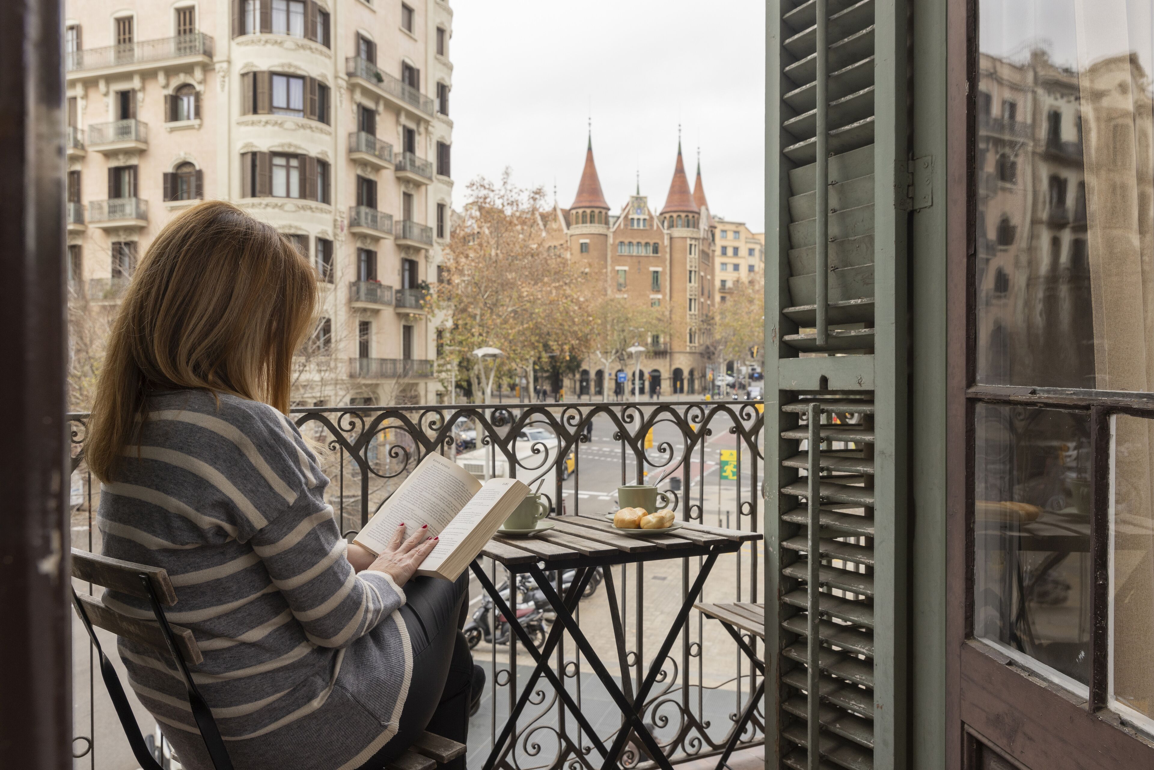 Una persona sentada en un balcón con un libro, una taza y un plato de pasteles.