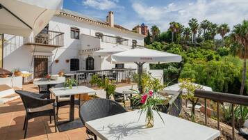 Un patio al aire libre con mesas y sillas, un paraguas blanco y un jarrón con flores.