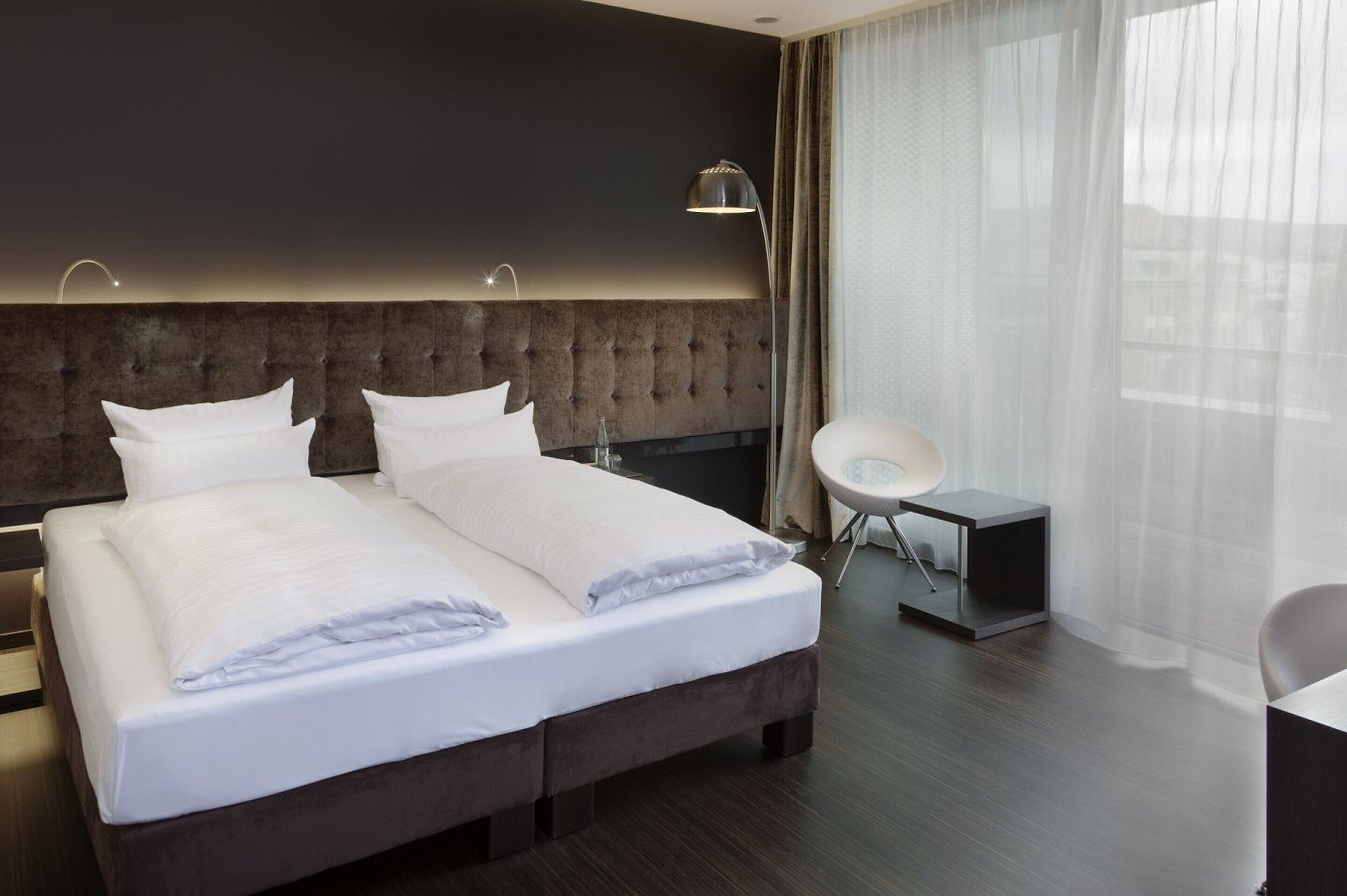 A modern hotel room with a large bed, a bedside table, a chair, and a large window with sheer curtains.
