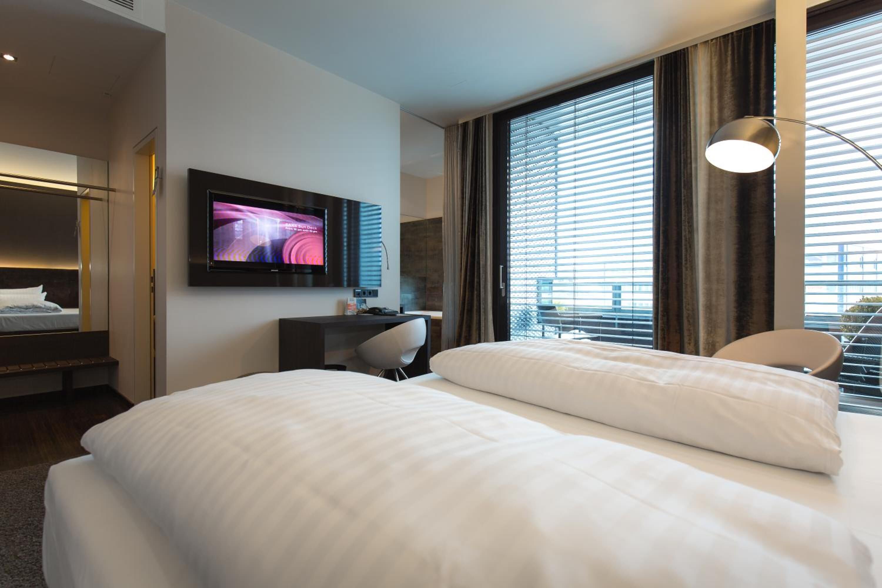A hotel room with a bed, a desk, a chair, a TV, and a window with blinds.