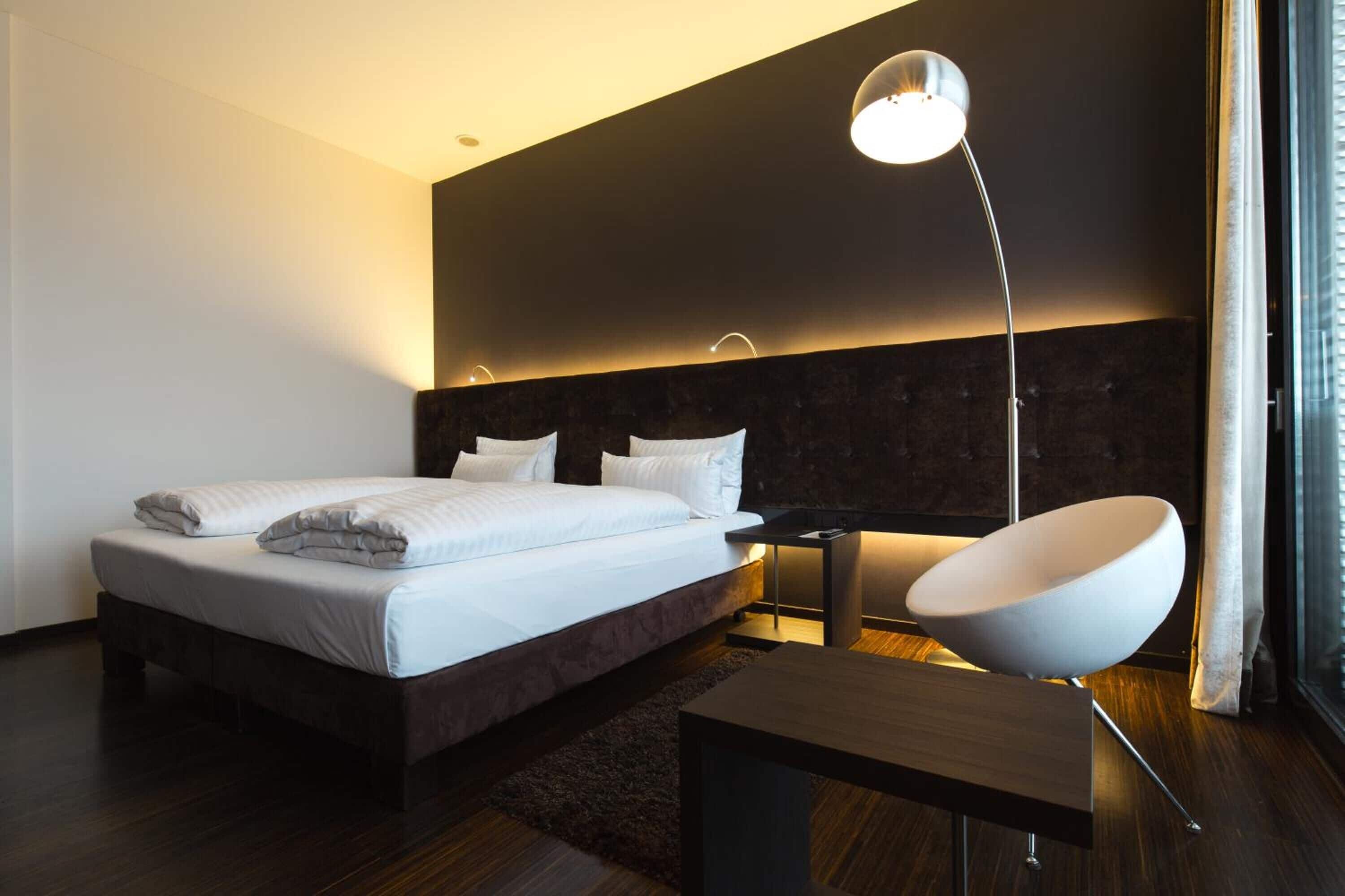 A modern hotel room with a large bed, a bedside table, a modern chair, and a floor lamp.