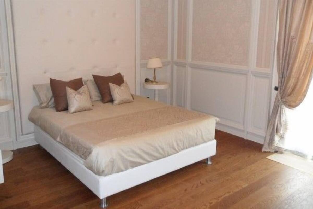 A bedroom with a white bed, beige bedding, and a small bedside table with a lamp.