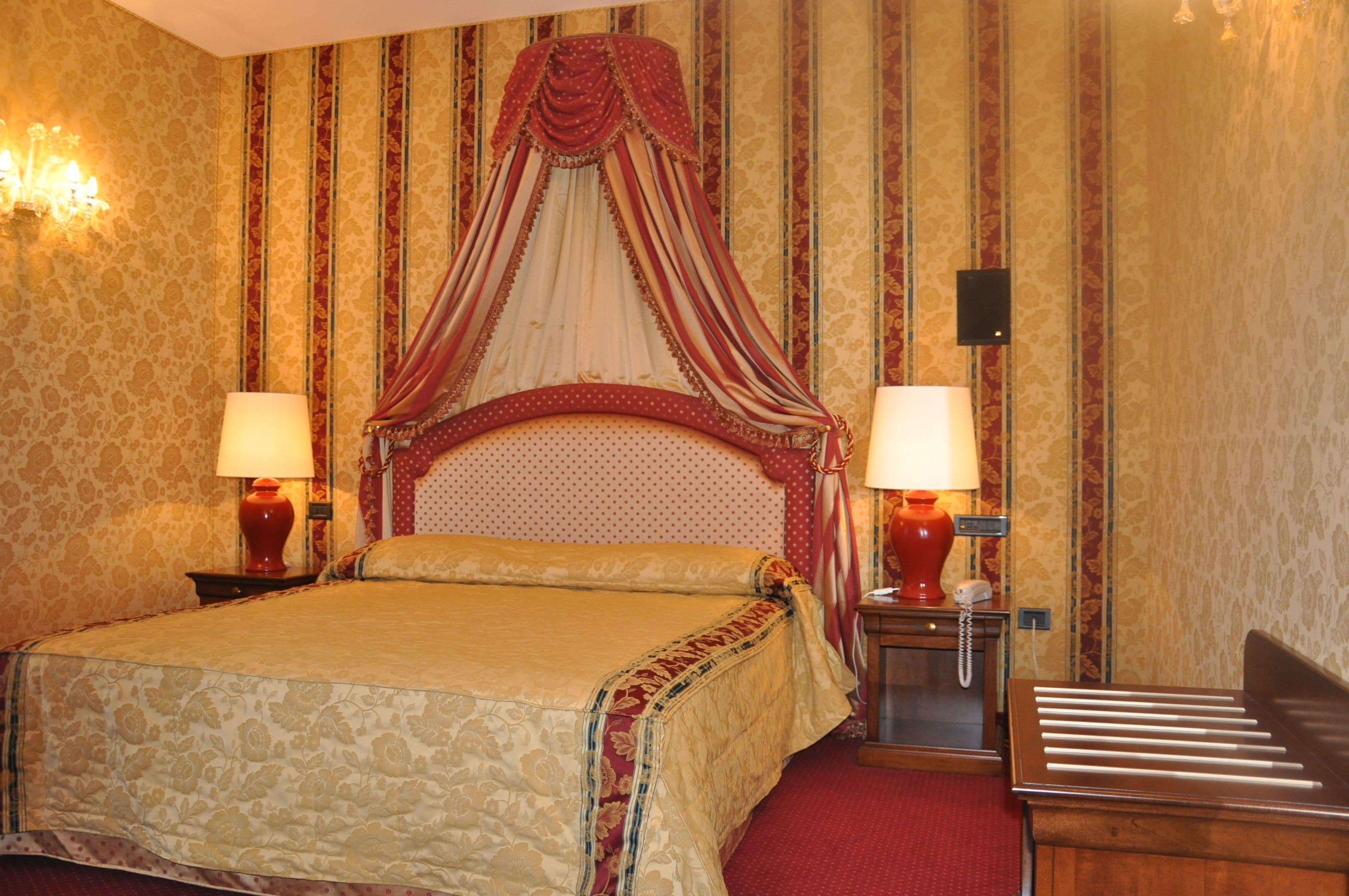 Habitación de hotel con cama con dosel, dos lámparas de noche, un teléfono y suelo cubierto de alfombra roja.