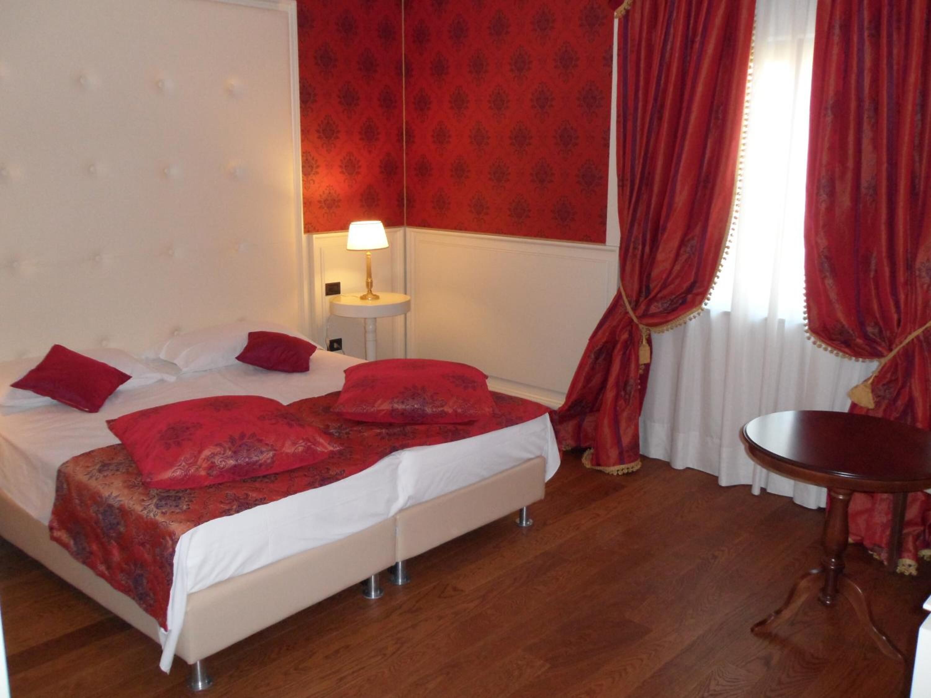 Une chambre d’hôtel avec un lit, une table de chevet avec une lampe, une chaise et une fenêtre avec des rideaux rouges.