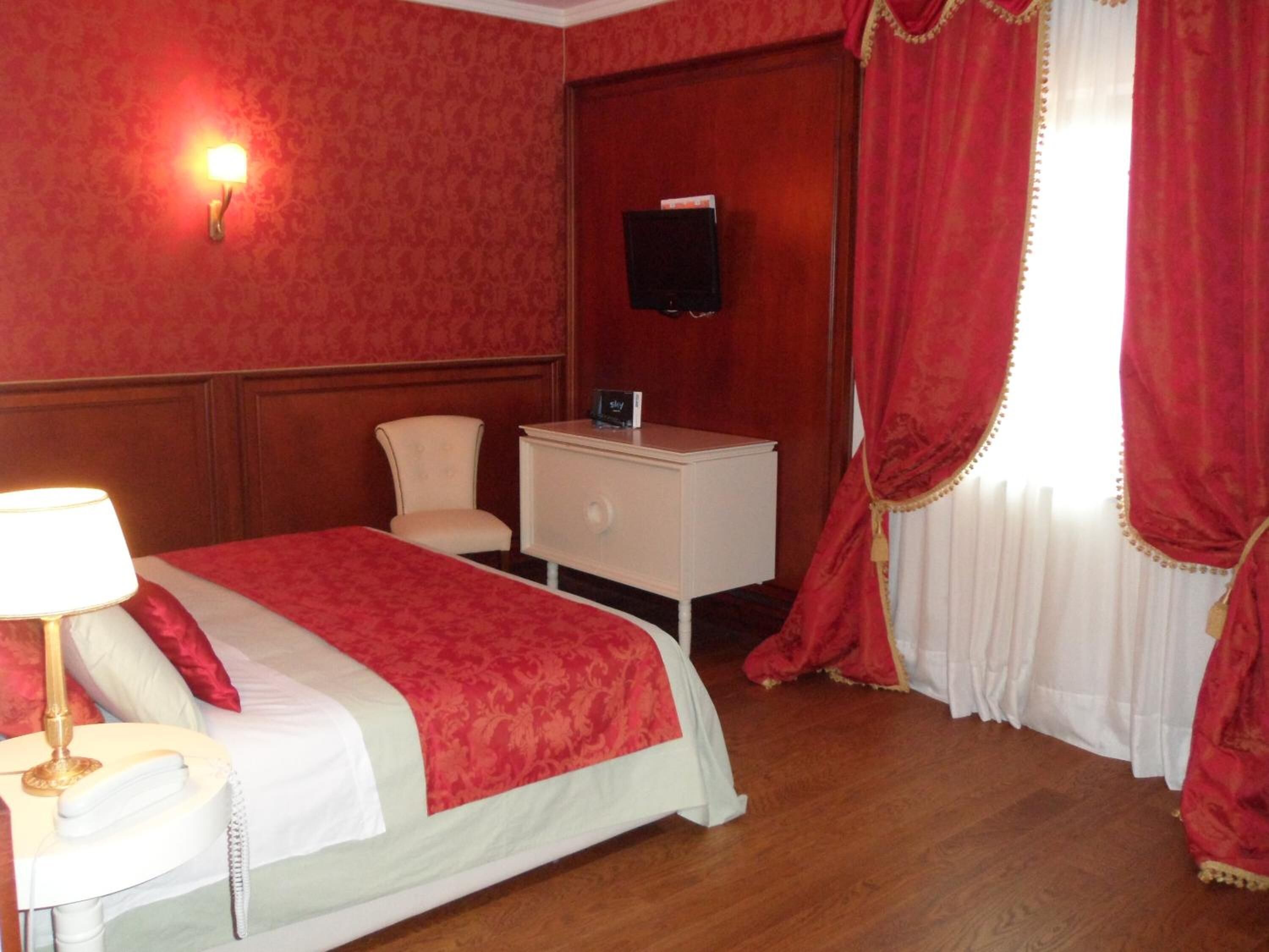 Una camera d'albergo con un letto, un comodino con una lampada, una TV a parete e una finestra con tende rosse.