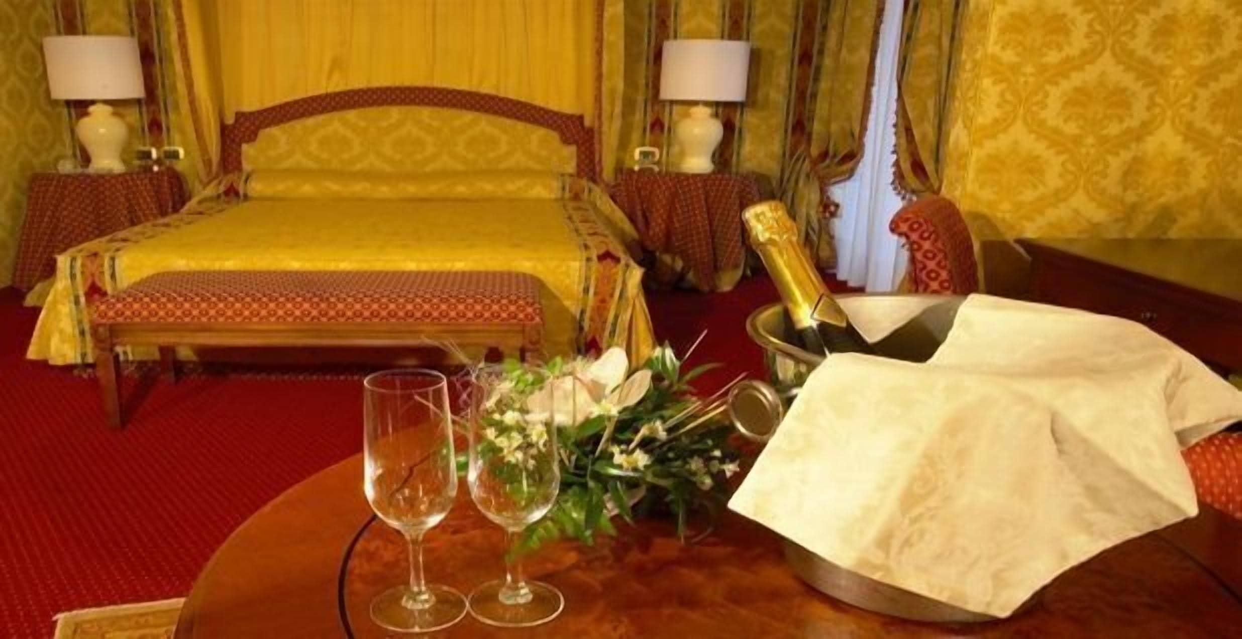 A hotel room with a bed, two bedside tables with lamps, a round table with a bottle of champagne, two wine glasses, and a bouquet of flowers.