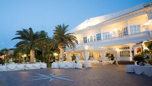 Een statig hotel met een ruim buitengebied, palmbomen en stijlvol ingerichte witte tafels voor een evenement.