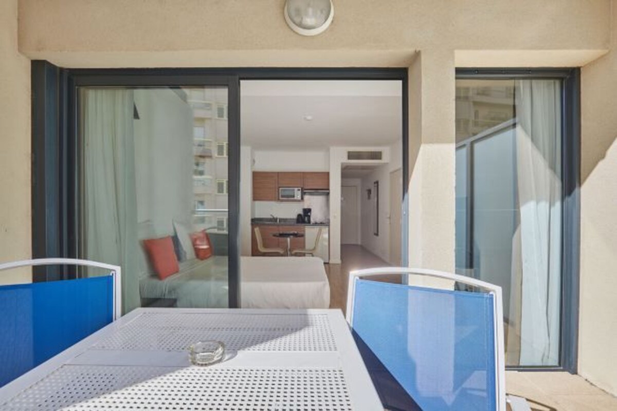A balcony with a table and chairs, a sliding glass door leading to a room with a bed and a kitchen area.