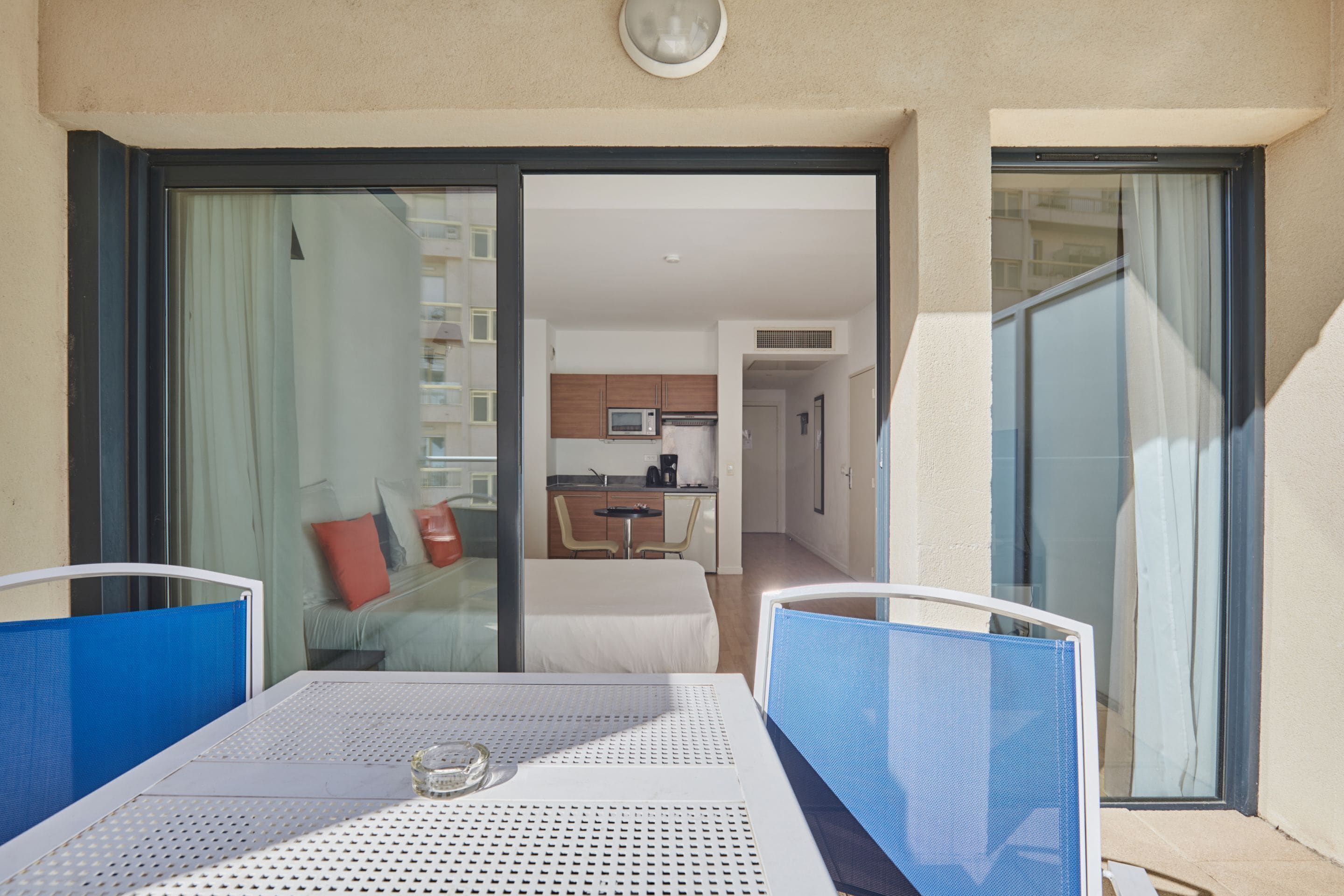 A balcony with a table and chairs, a sliding glass door, and a glimpse of a kitchen and living area inside.