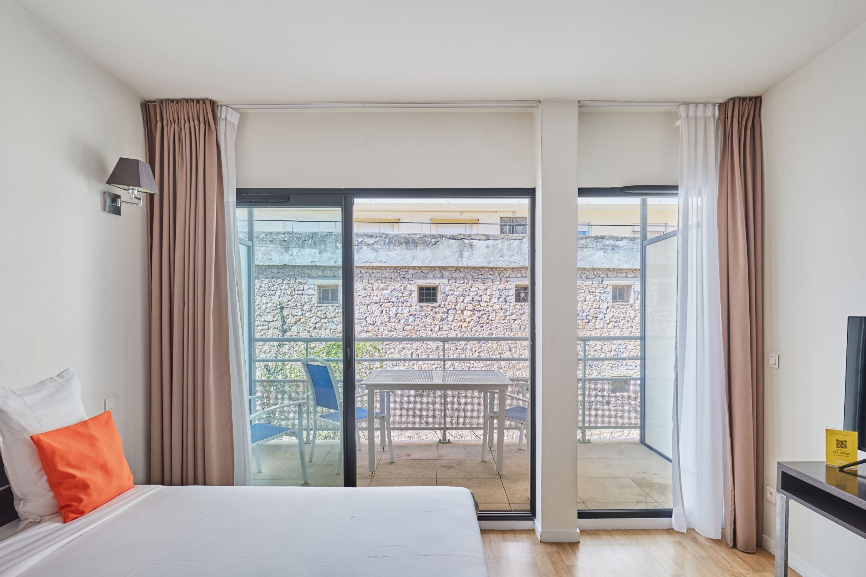 Quarto de hotel com cama, uma mesa e vista para um edifício de pedra através de uma porta de vidro.