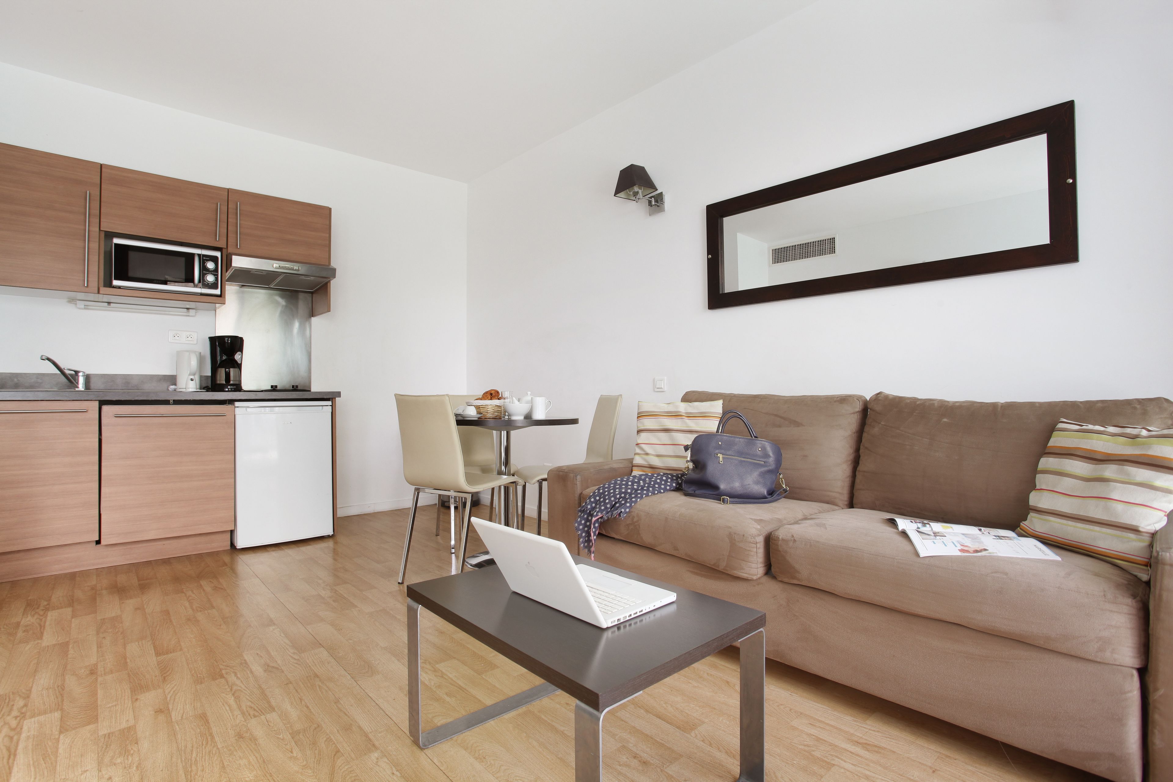 Een moderne woonkamer met een bank, een salontafel met een laptop, een eethoek met stoelen, en een keuken met een magnetron en koffiezetapparaat.