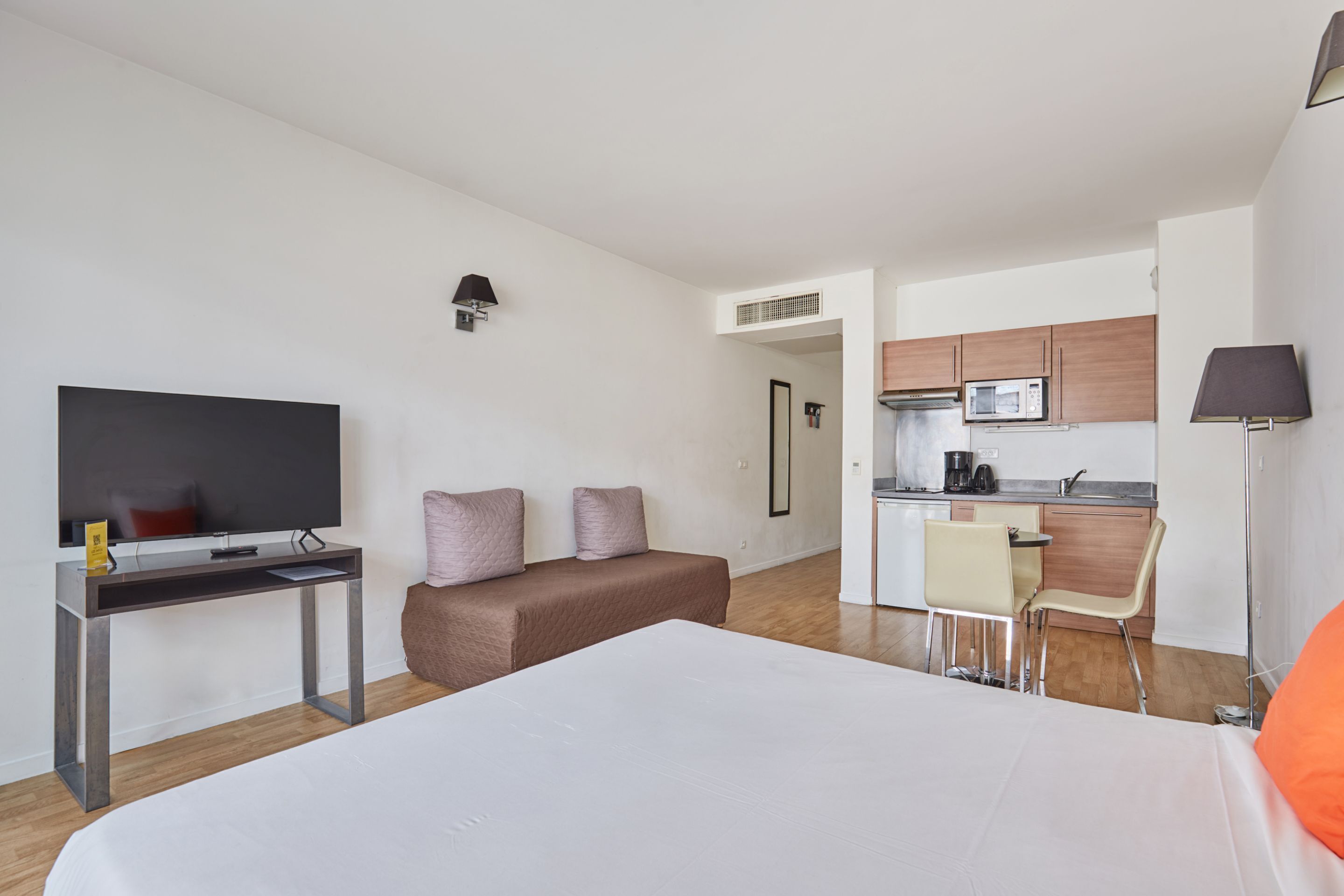 Una habitación de hotel moderna con cama, escritorio con televisor de pantalla plana, kitchenette y zona de estar con una silla y una mesa.