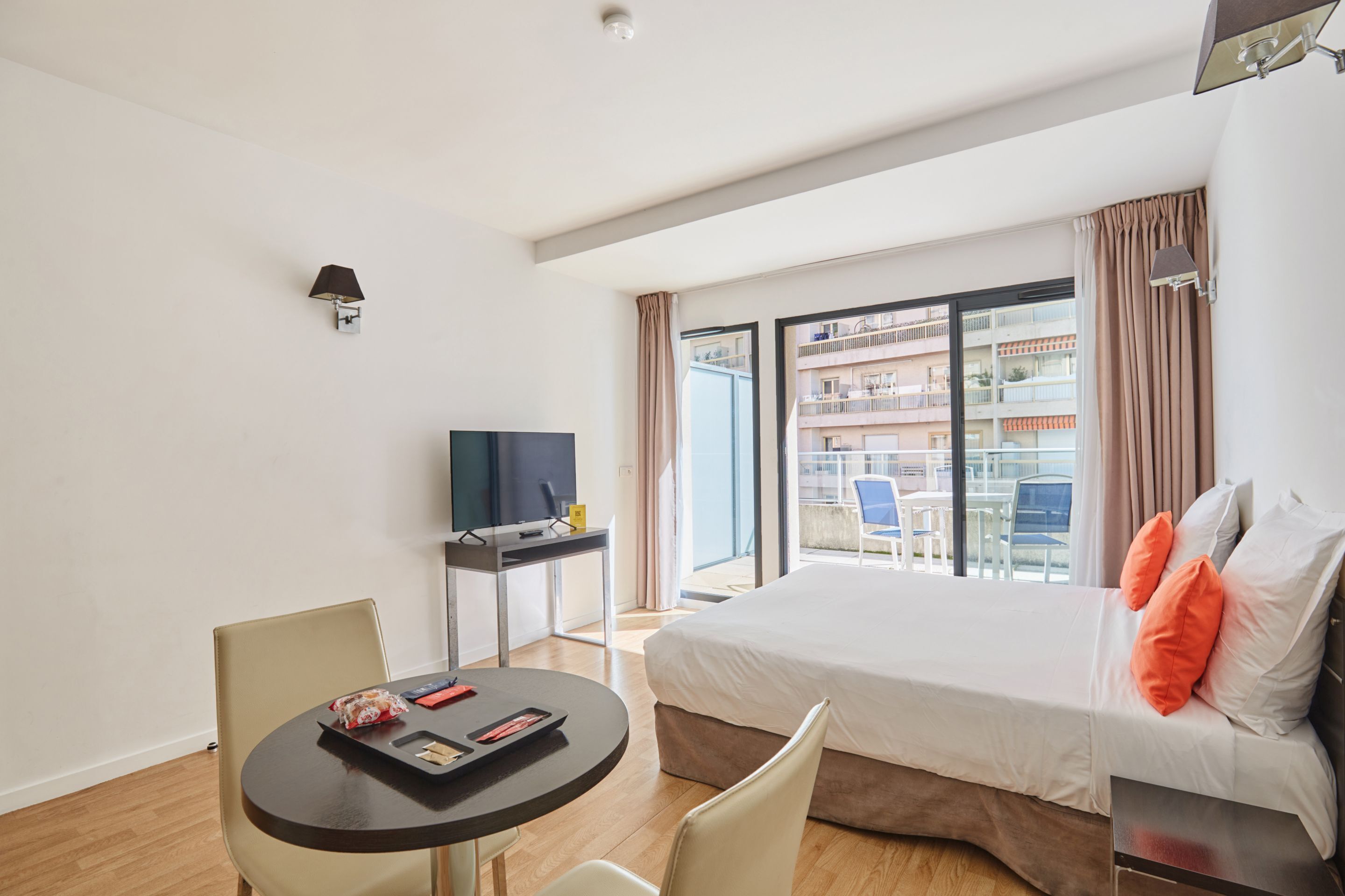 A hotel room with a bed, a desk, a chair, a TV, and a balcony with a view of other buildings.