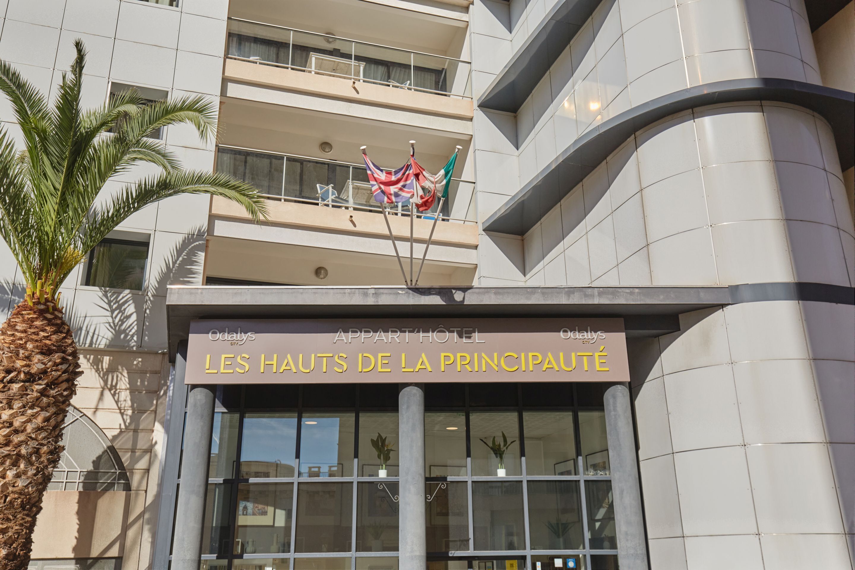 A hotel entrance with a sign reading 'LES HAUTS DE LA PRINCIPAUTÉ,' balconies, and a palm tree.
