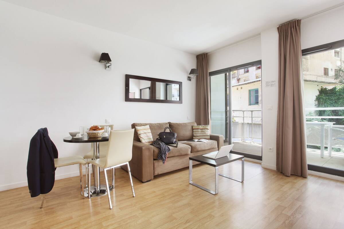 Un salon moderne avec un canapé, une table à manger et des chaises, un ordinateur portable, et une vue sur le balcon extérieur.