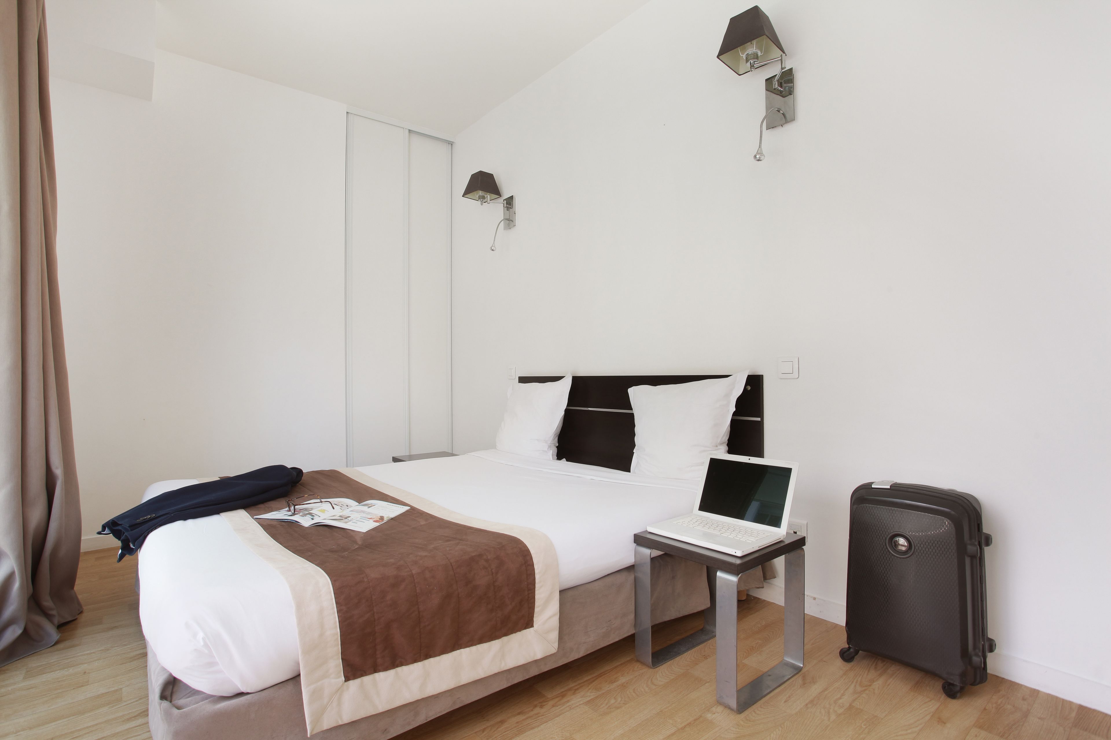 Una habitación de hotel con cama, escritorio, computadora portátil, maleta y lámpara de pared.