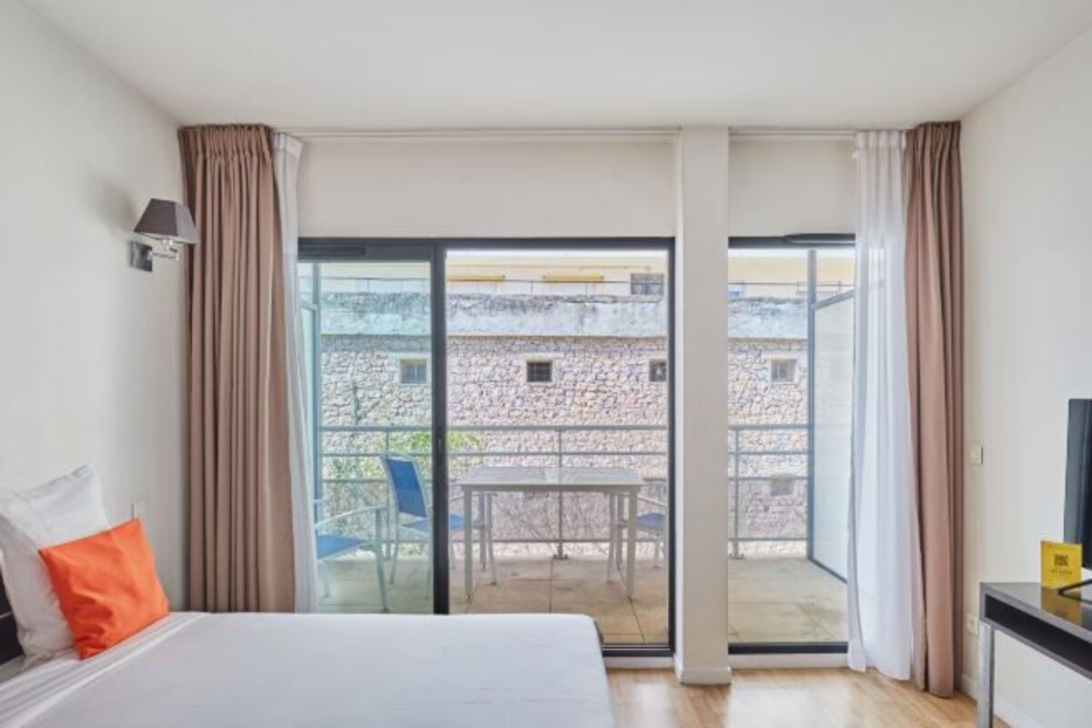 A hotel room with a bed, a small table, and a view through a sliding glass door to an outdoor area.