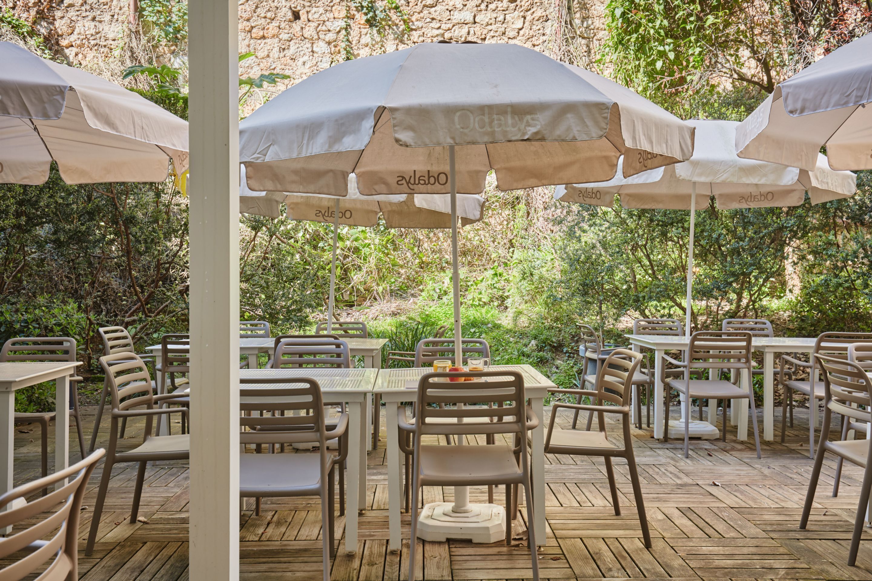 Un espacio de comedor al aire libre con mesas y sillas, cubierto por sombrillas, frente a un muro de piedra y follaje.