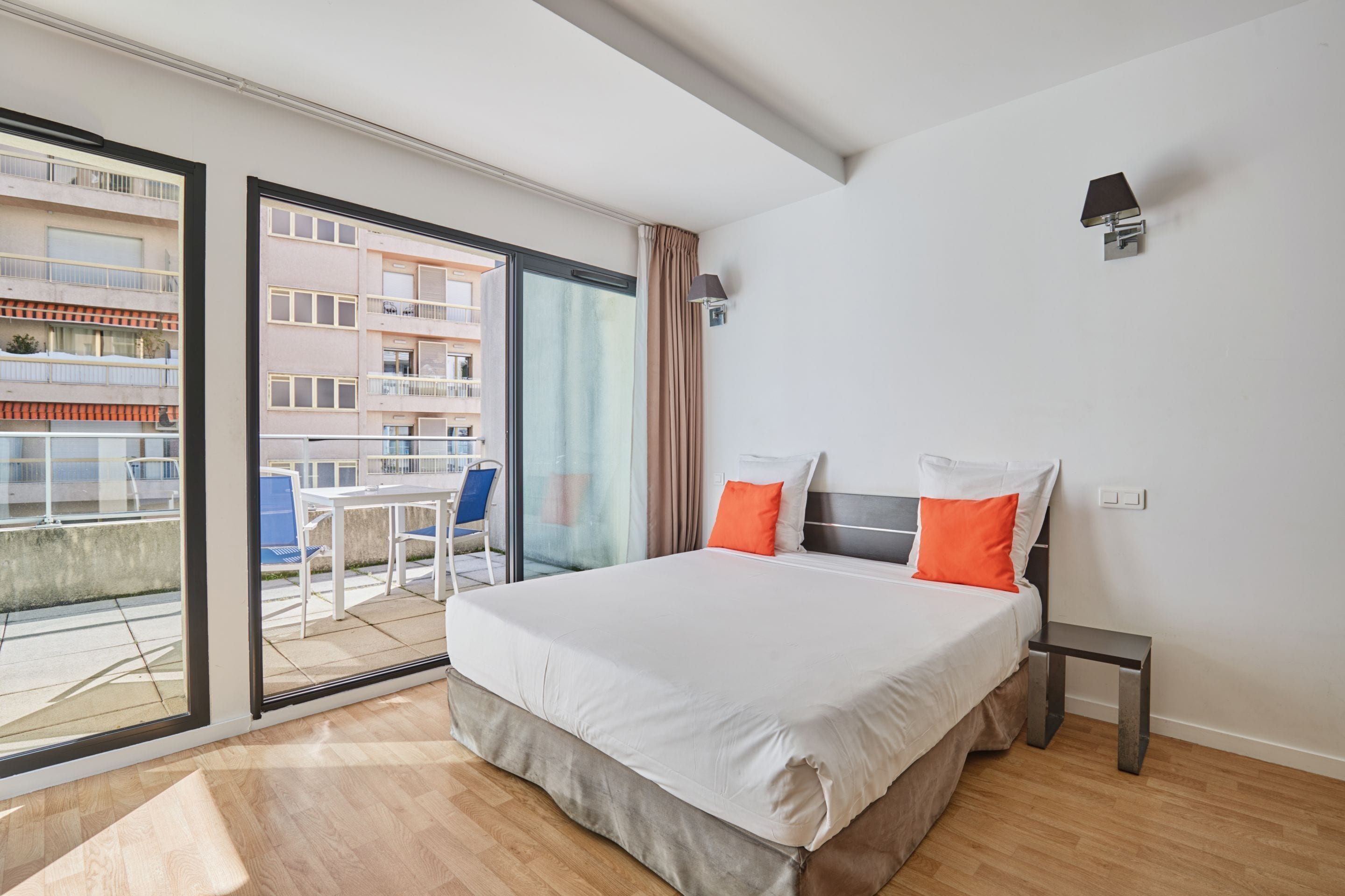 Une chambre d’hôtel avec un grand lit, une petite table, une chaise et un balcon donnant sur d’autres immeubles.