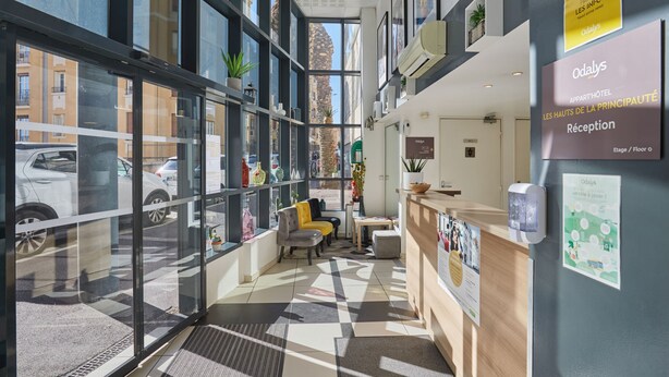 A hotel reception area with a check-in counter, seating area, and large windows offering views of the street. - Appart'hôtel Odalys City - Monaco Les Hauts de la Principauté (Beausoleil)