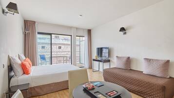Ein Hotelzimmer mit Bett, einer Couch, einem kleinen Tisch mit Snacks, einem Fernseher und Blick auf Gebäude durch große Fenster.