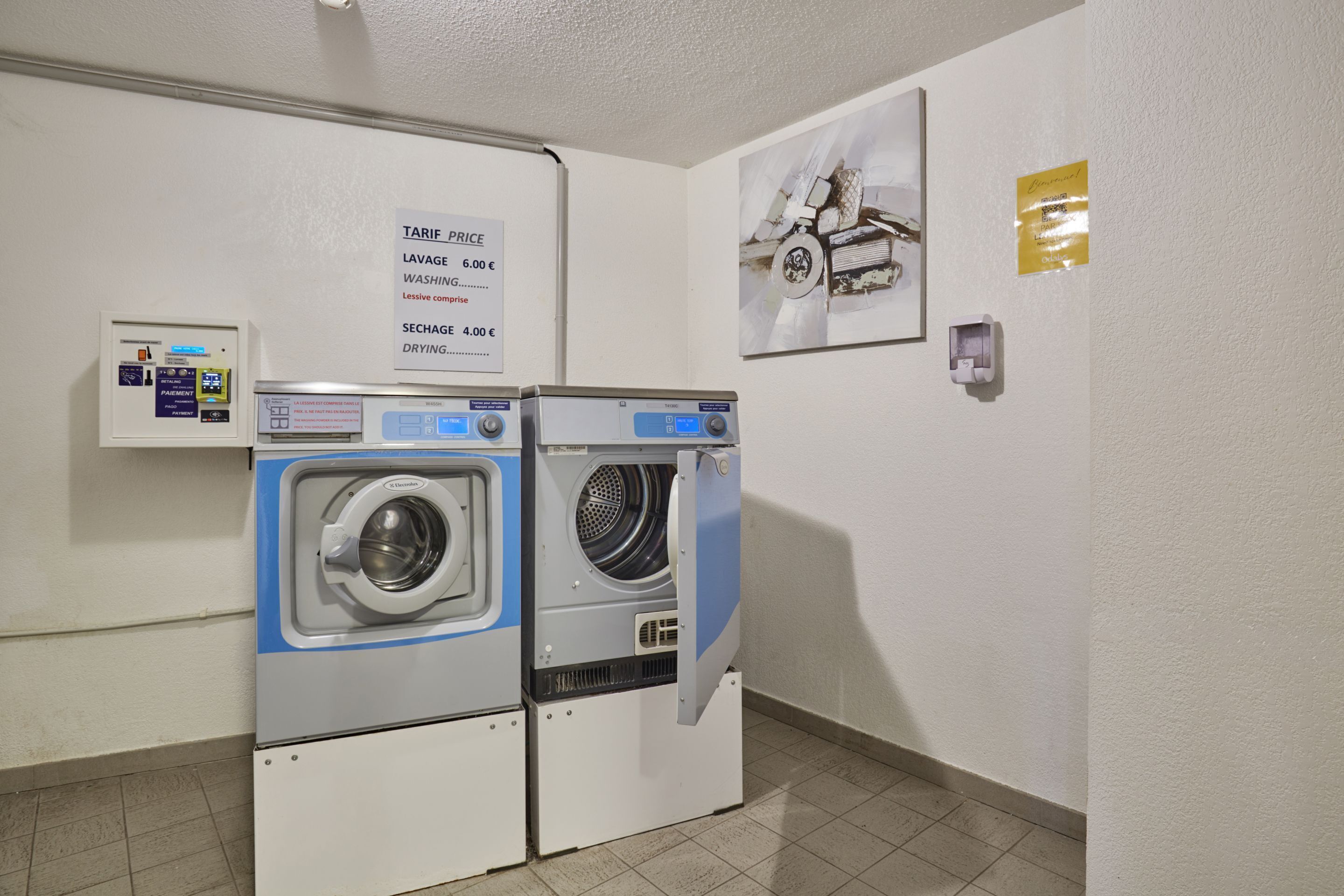 A laundry area with two front-loading washing machines, a price sign, a small control panel, and a framed picture on the wall.