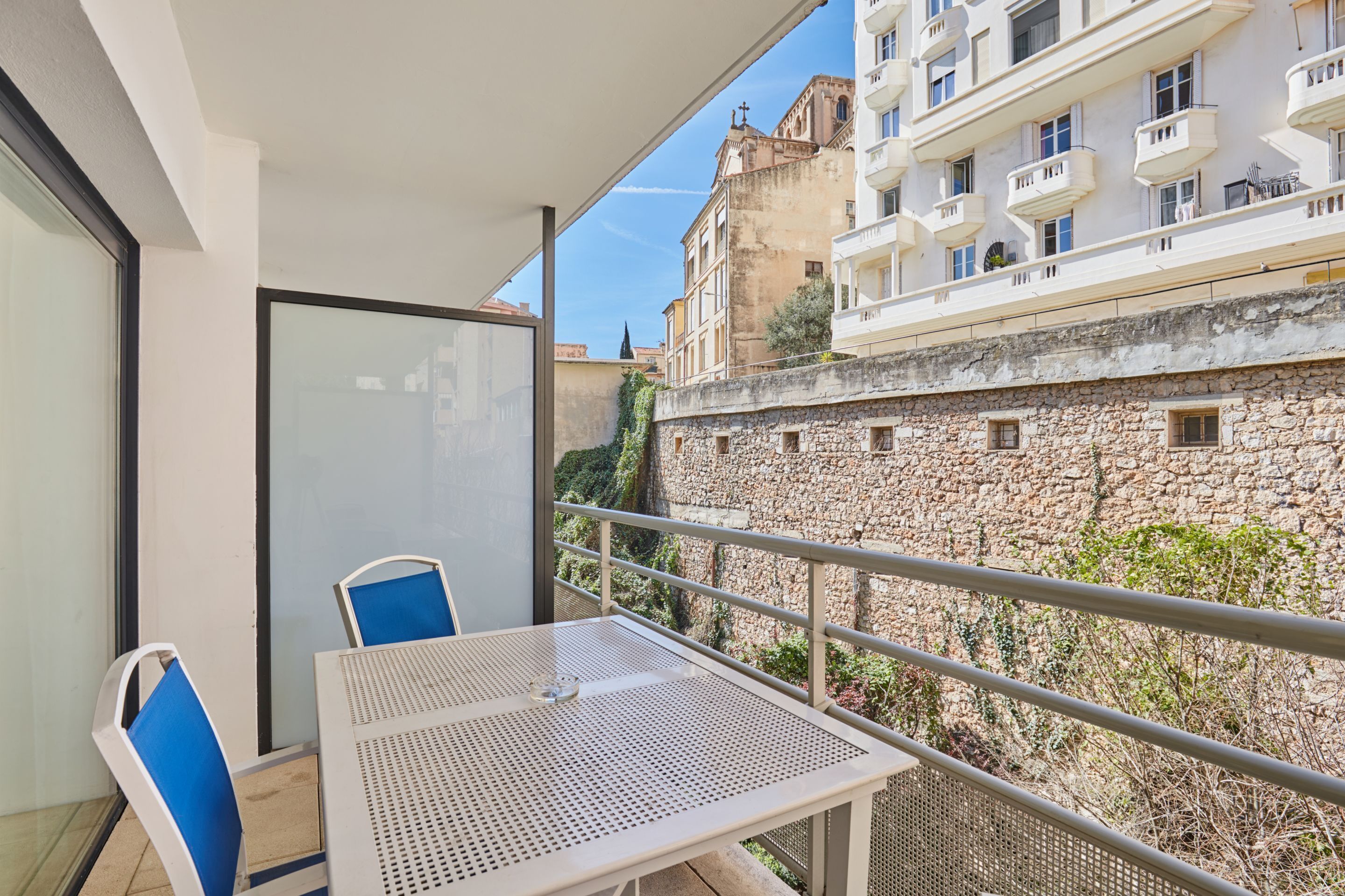 Un balcón con mesa y sillas, con vista a un muro de piedra y edificios.