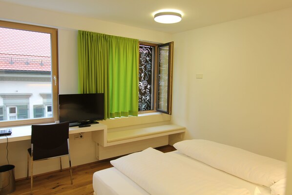 Un dormitorio con una cama, un escritorio con silla, un televisor y una ventana con cortinas verdes.