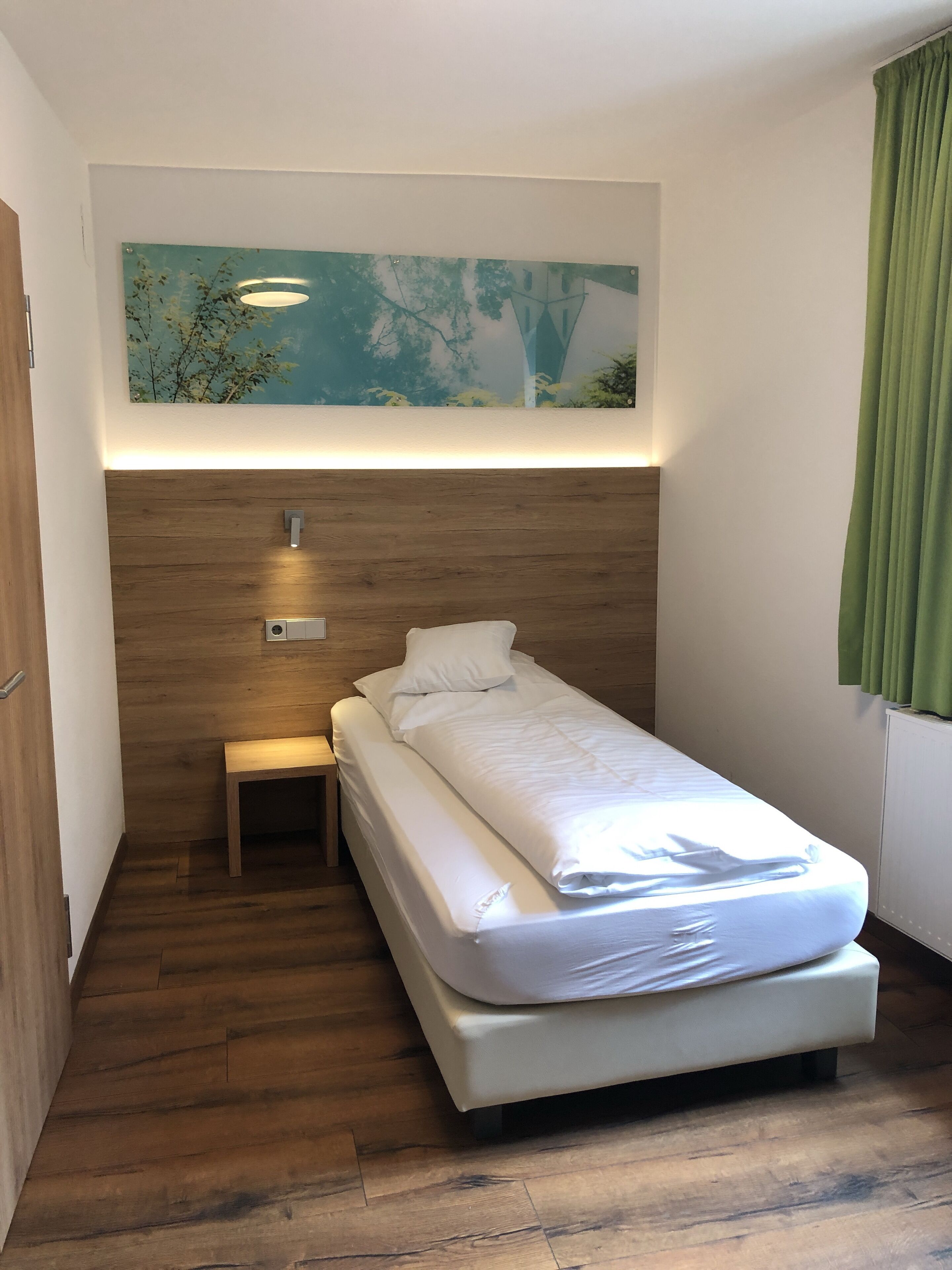 A small, neatly arranged bedroom with a single bed, a small bedside table, a wall-mounted light, and a painting above the bed.